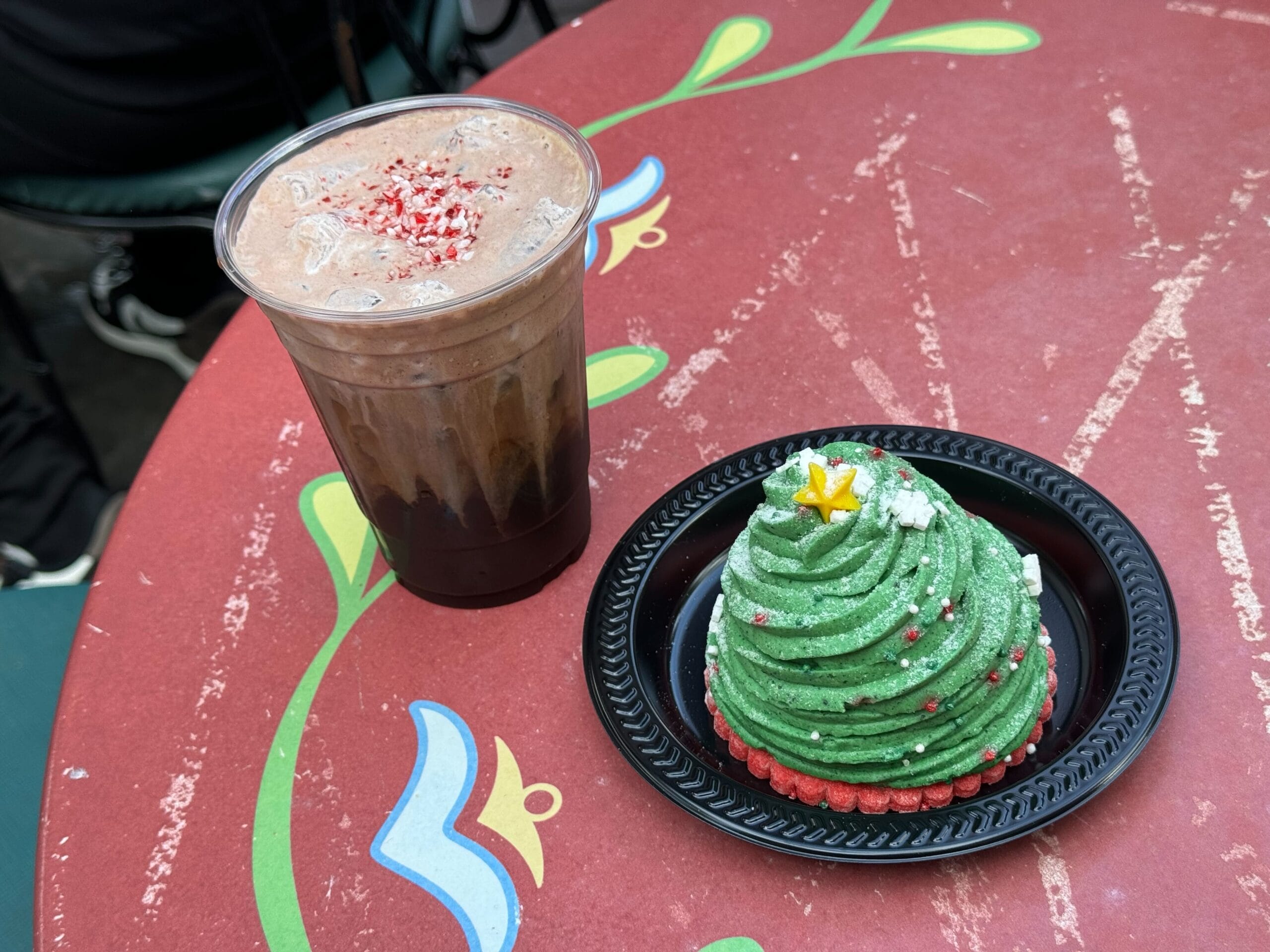 A Peppermint Mocha Cold Brew sits next to a green, tree-shaped dessert, evoking Christmas cheer on a red table with floral designs.