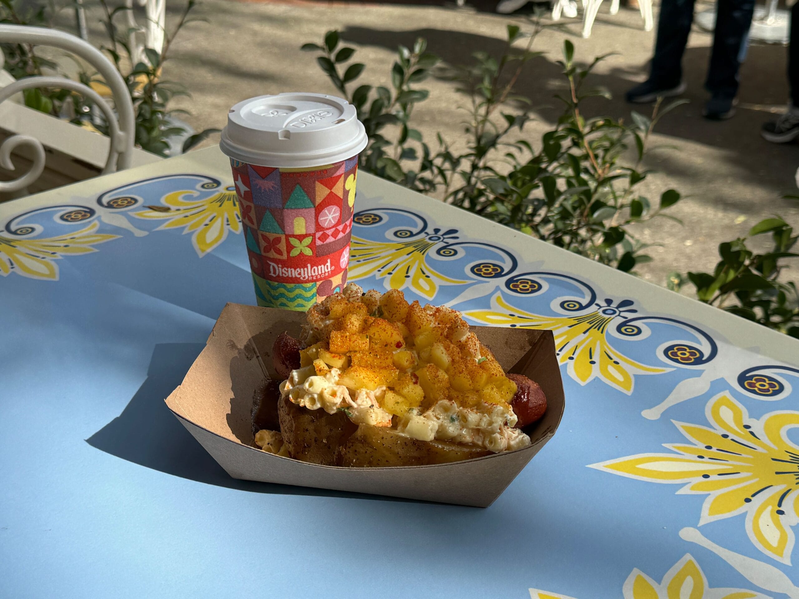 At the Refreshment Corner, enjoy a hot dog topped with creamy macaroni and cheese, presented in a paper tray alongside a lidded disposable cup on a festively adorned outdoor table.