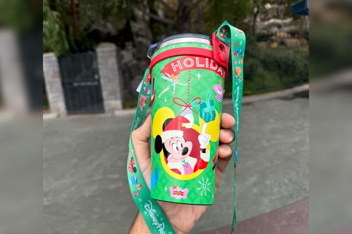 A hand holds a festive green, holiday stainless steel tumbler featuring a cartoon character in joyful holiday attire, complete with a matching lanyard attached.