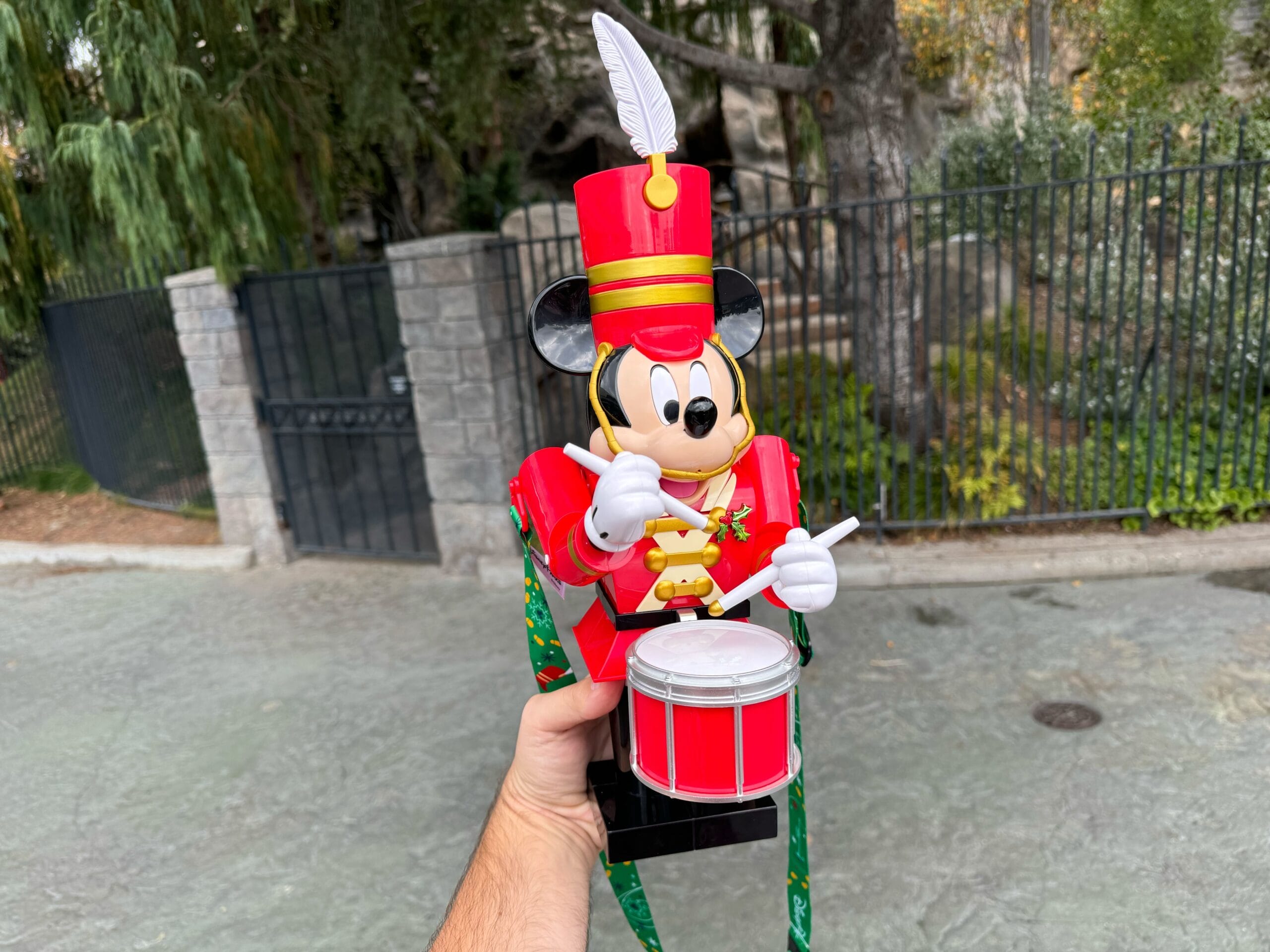 A person holds a Mickey Toy Soldier, a drumming cartoon mouse in a red uniform with a feathered hat, set against an outdoor backdrop with trees and a stone fence.