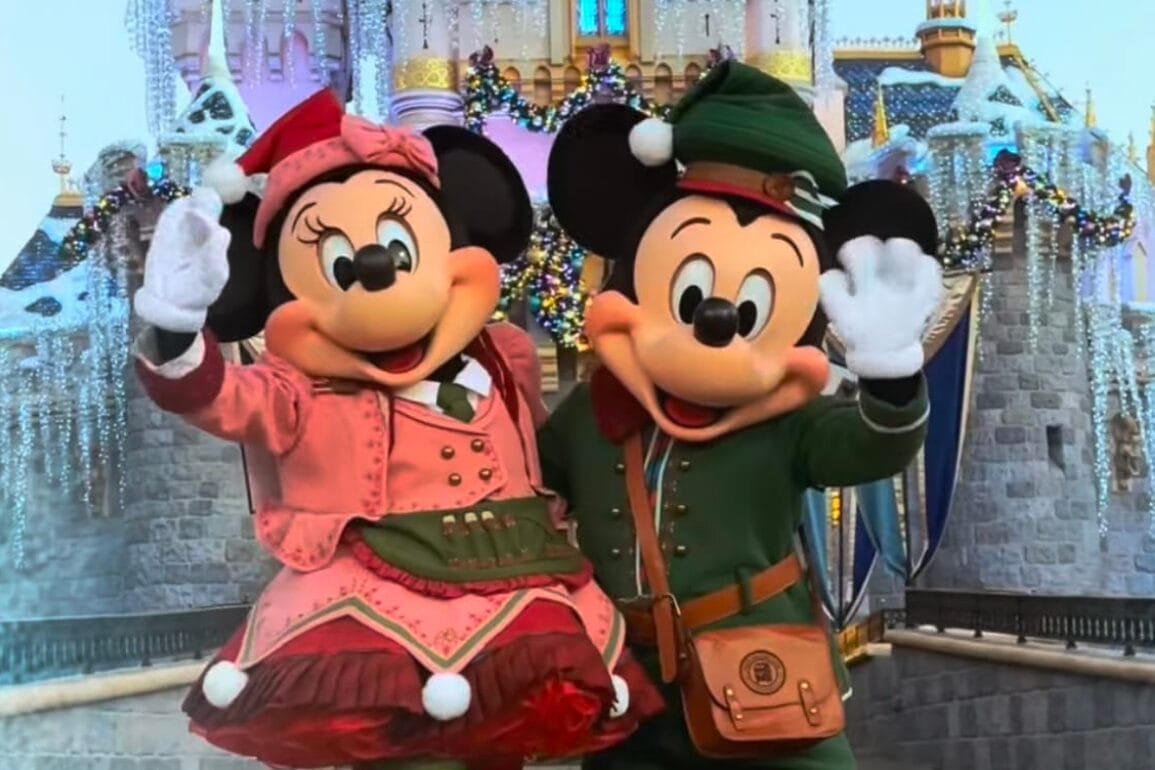 Two costumed characters in festive attire wave joyfully in front of a beautifully decorated castle at Disneyland.