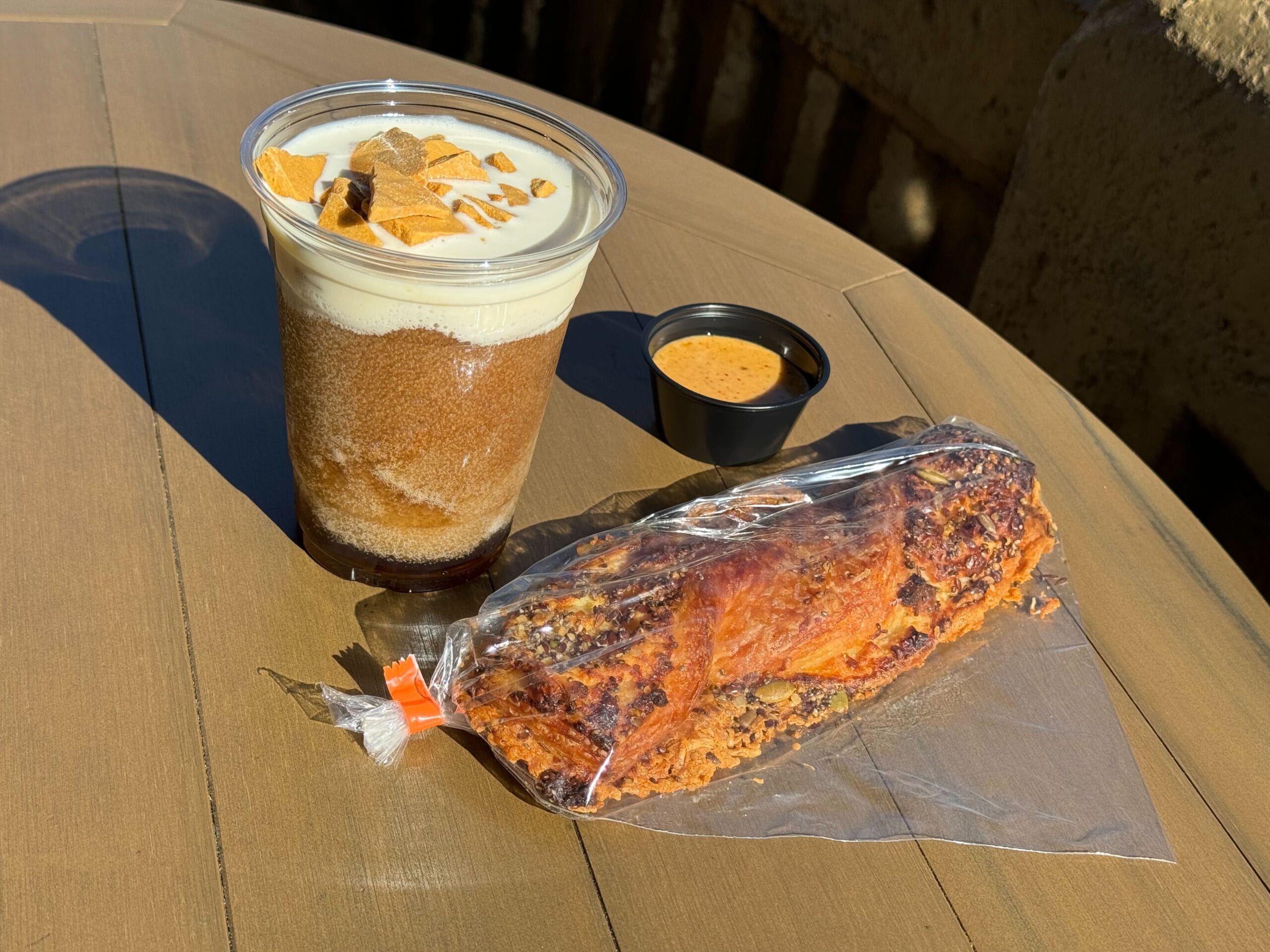 Iced beverage topped with cream and chips, a side of dipping sauce, and a wrapped pastry on a wooden table.