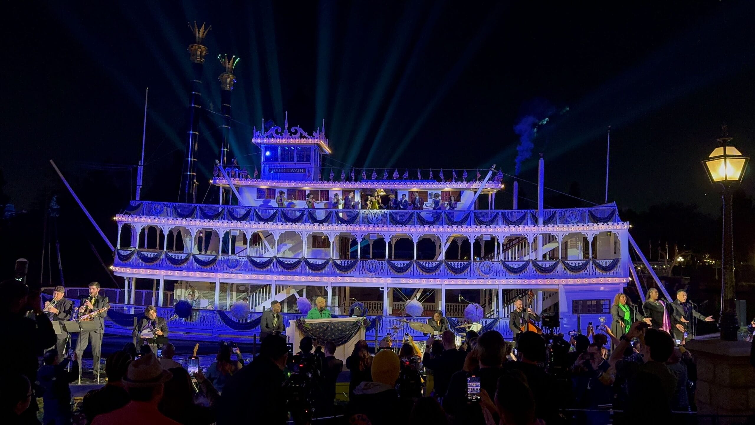 A large, illuminated riverboat with three decks is adorned with lights and decorations, set against a night sky. People are gathered around taking photos.