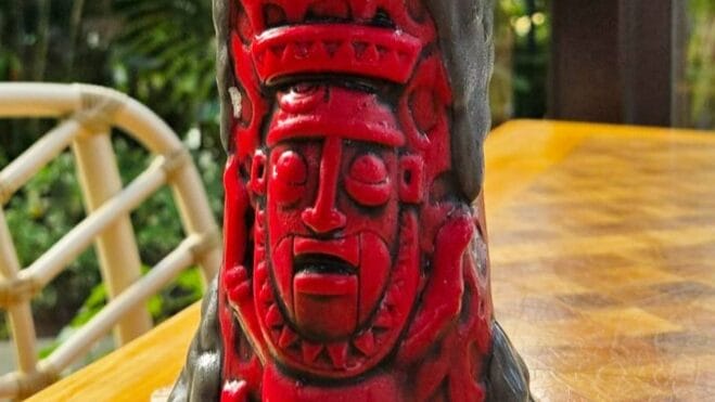 Red and black carved tiki mug on a wooden table with greenery in the background.