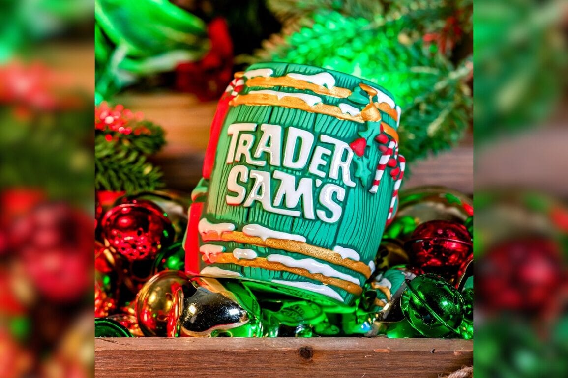 Colorful mug with "Trader Sam's" text sits among festive greenery and shiny ornaments in red, gold, and green hues.