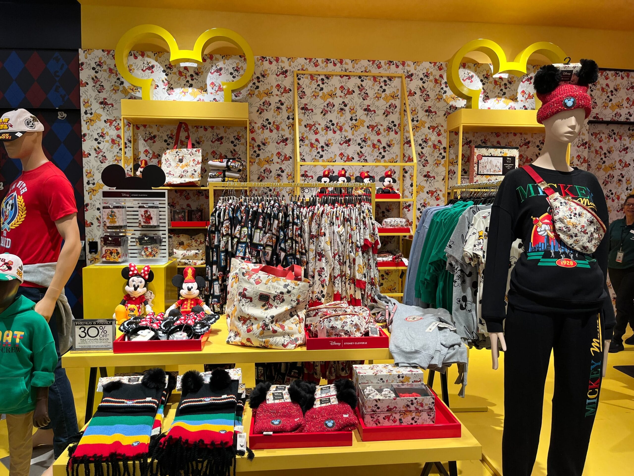 Store display with Disney-themed apparel and accessories, including hoodies, hats, and backpacks. Mannequins and shelves showcase Mickey Mouse designs. Bright yellow decor with floral wallpaper.