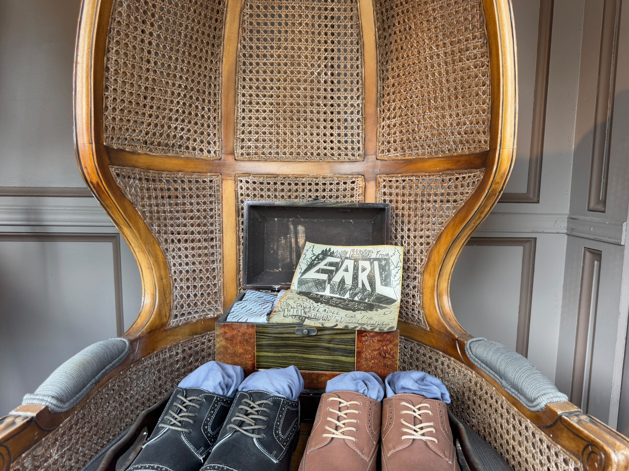 Two pairs of shoes and a box with a folded cloth sit inside a woven chair. A bag labeled "EARL" leans against the box.