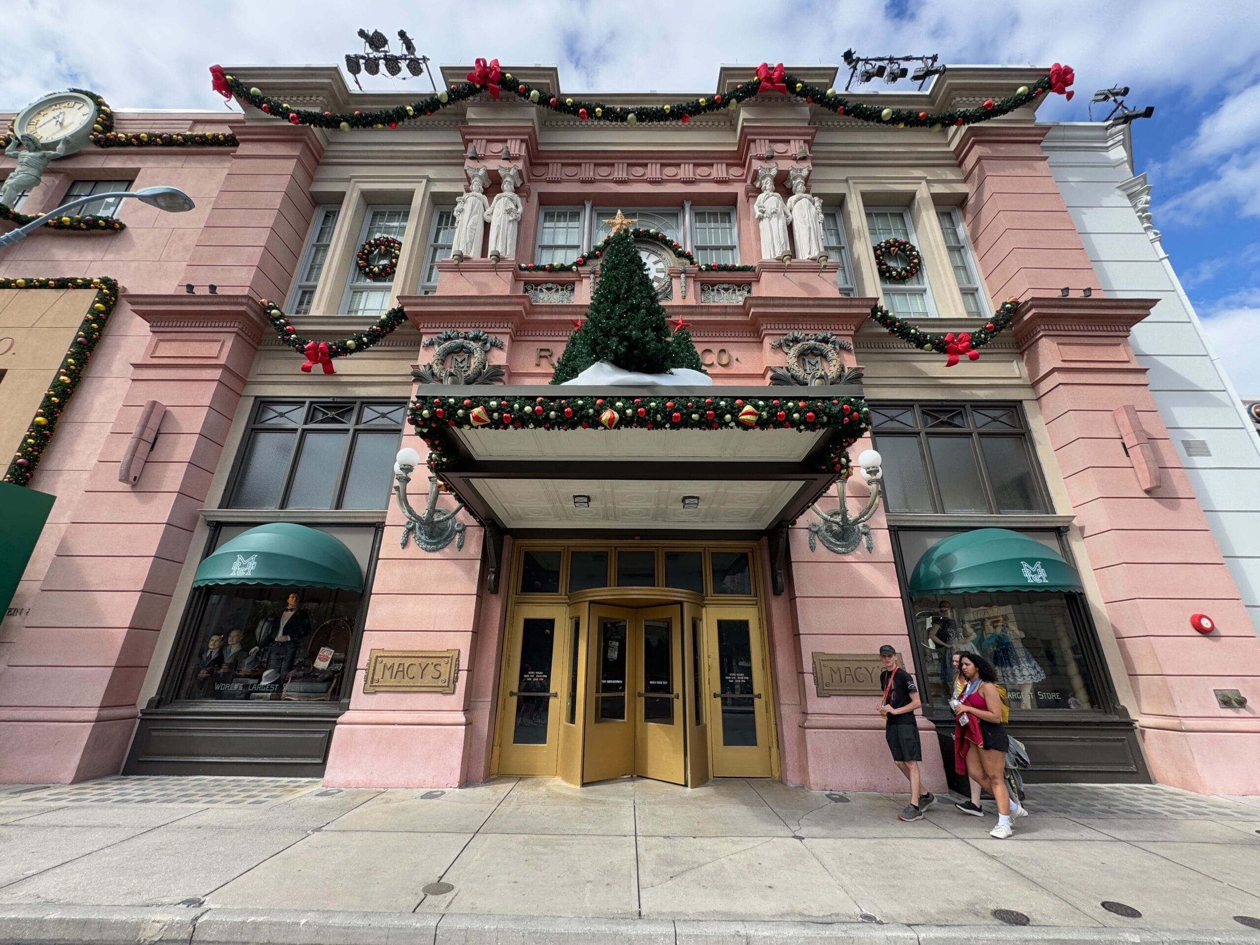 Facade of a pink building decorated with a Christmas tree and wreaths. Two people stand at the entrance under a vintage-style awning.