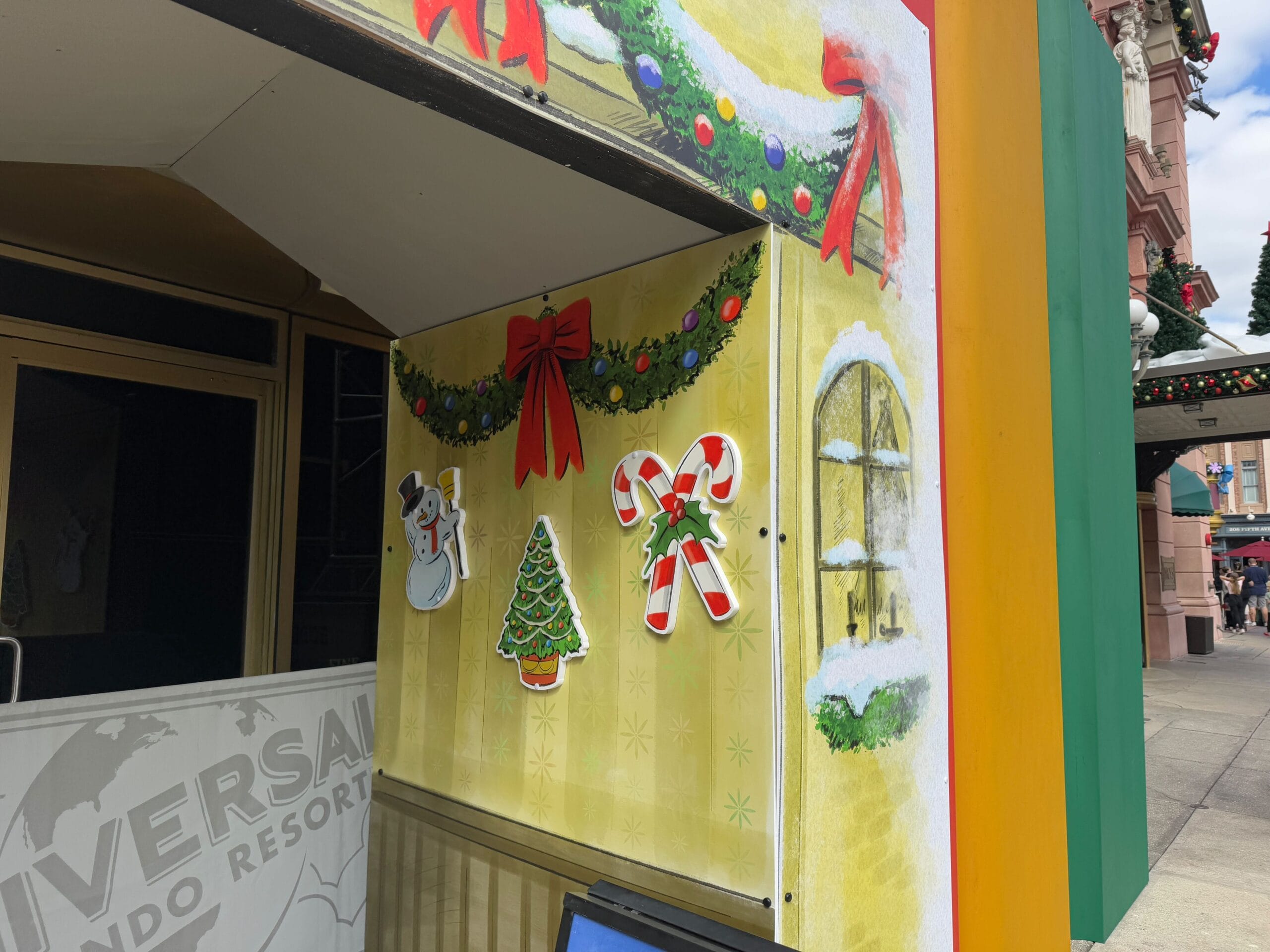 Holiday-themed decorations with snowflakes, a snowman, Christmas tree, candy canes, and greenery adorn a building corner.