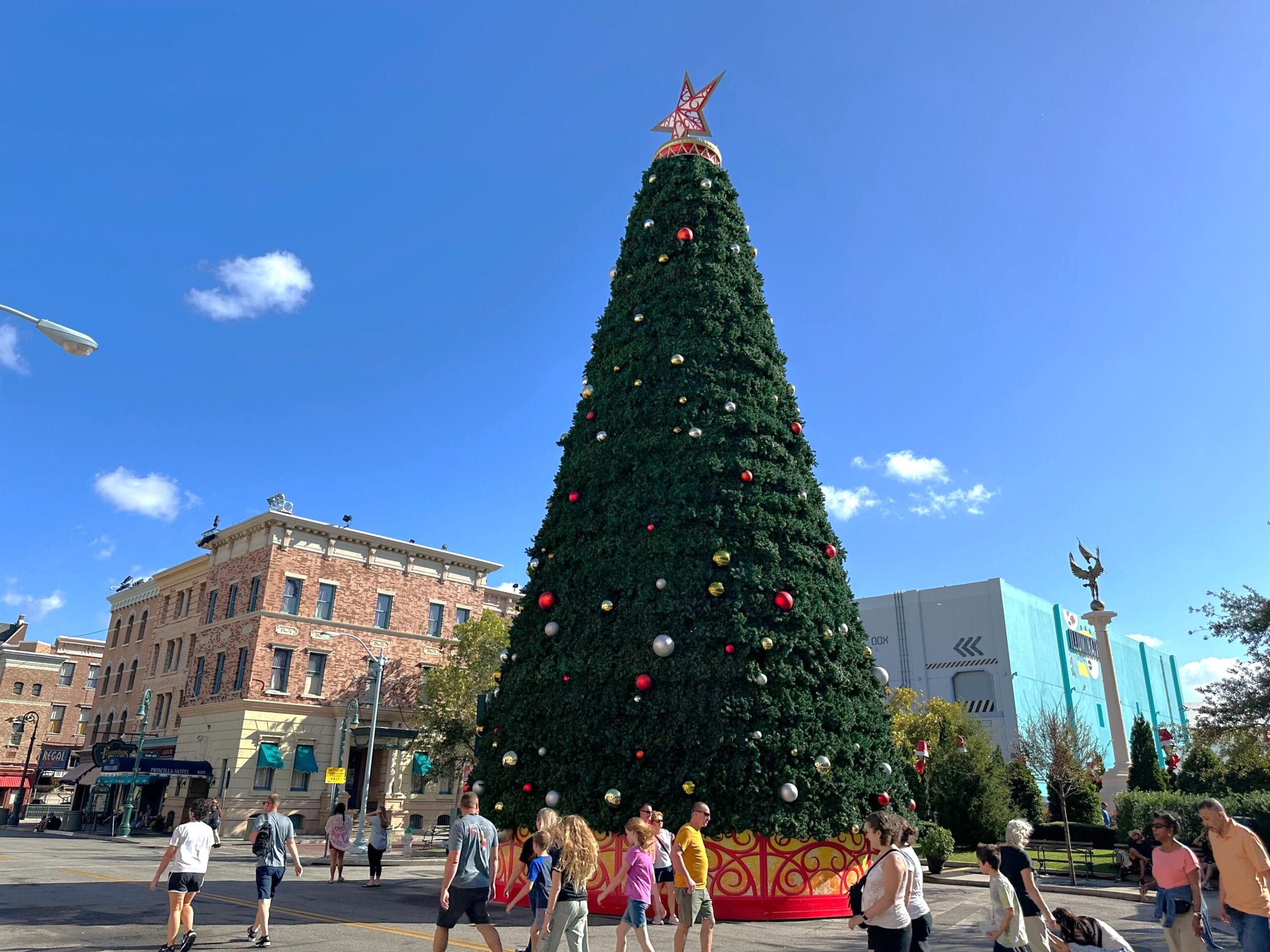 A tall Christmas tree adorned with ornaments stands in an urban plaza at Universal Studios Florida. People stroll beneath the clear blue sky, soaking in the festive atmosphere.