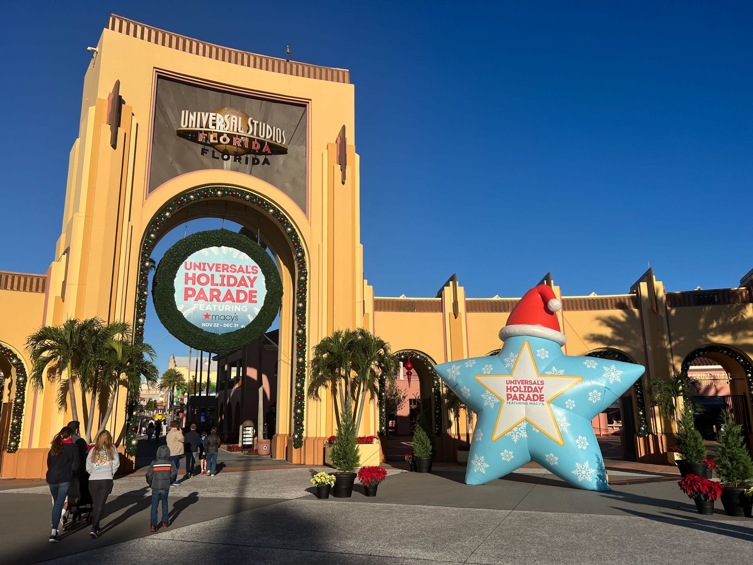 Entrance to Universal Studios Florida, featuring a large holiday parade sign and a star-shaped holiday decoration. People walk along the pathway under a clear blue sky.