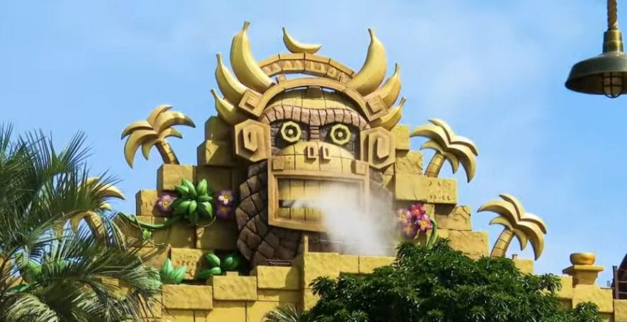 A sculpture resembling a golden monkey head with steam emerging from its mouth, reminiscent of Donkey Kong Country, is surrounded by greenery and decorative elements under a blue sky. This captivating piece could easily find a home in Universal Studios Japan's Super Nintendo World.