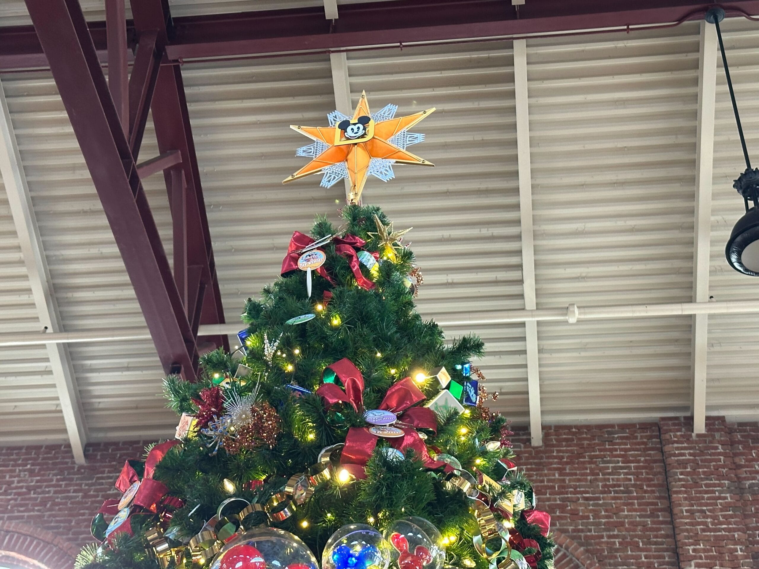 Christmas tree with a star topper decorated with lights, red bows, and ornaments. Indoor setting with a brick wall and metal beams in the background.