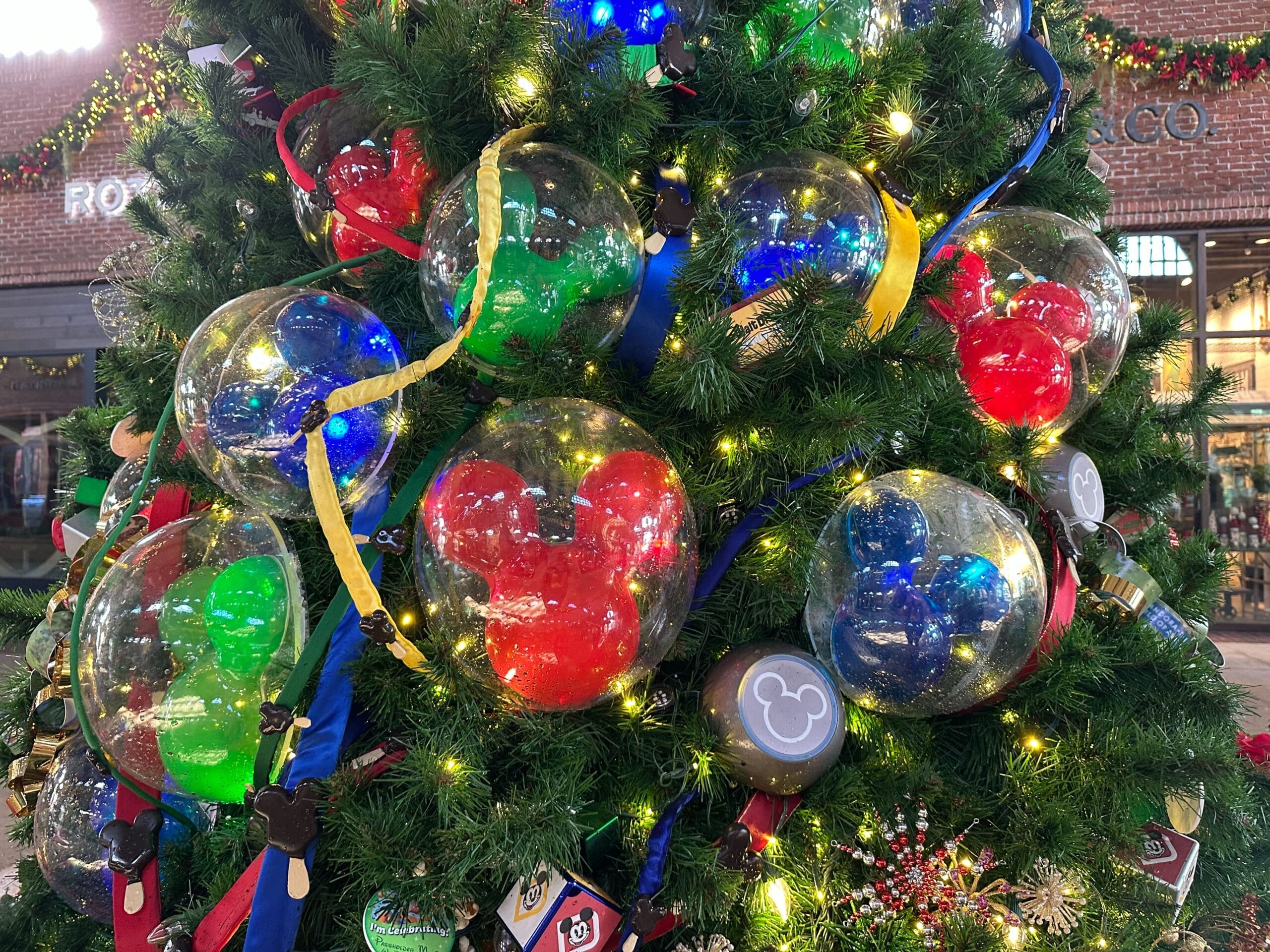 A Christmas tree decorated with clear ornaments filled with colored figures and ribbons, featuring Mickey Mouse shapes. Surrounding greenery is adorned with lights and festive decorations.