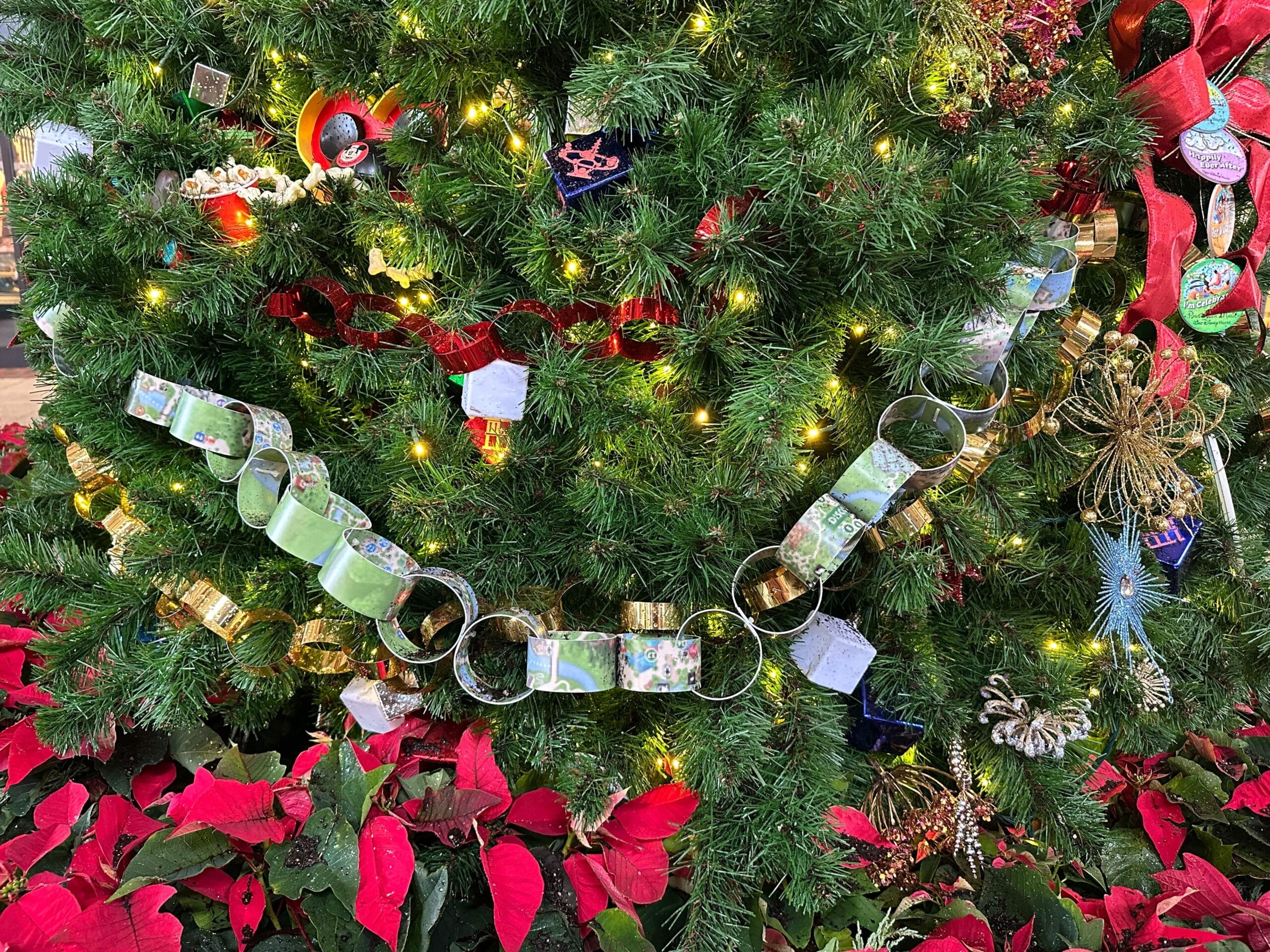 A decorated Christmas tree with a paper chain, ribbon, and ornaments. Red poinsettias surround the base.