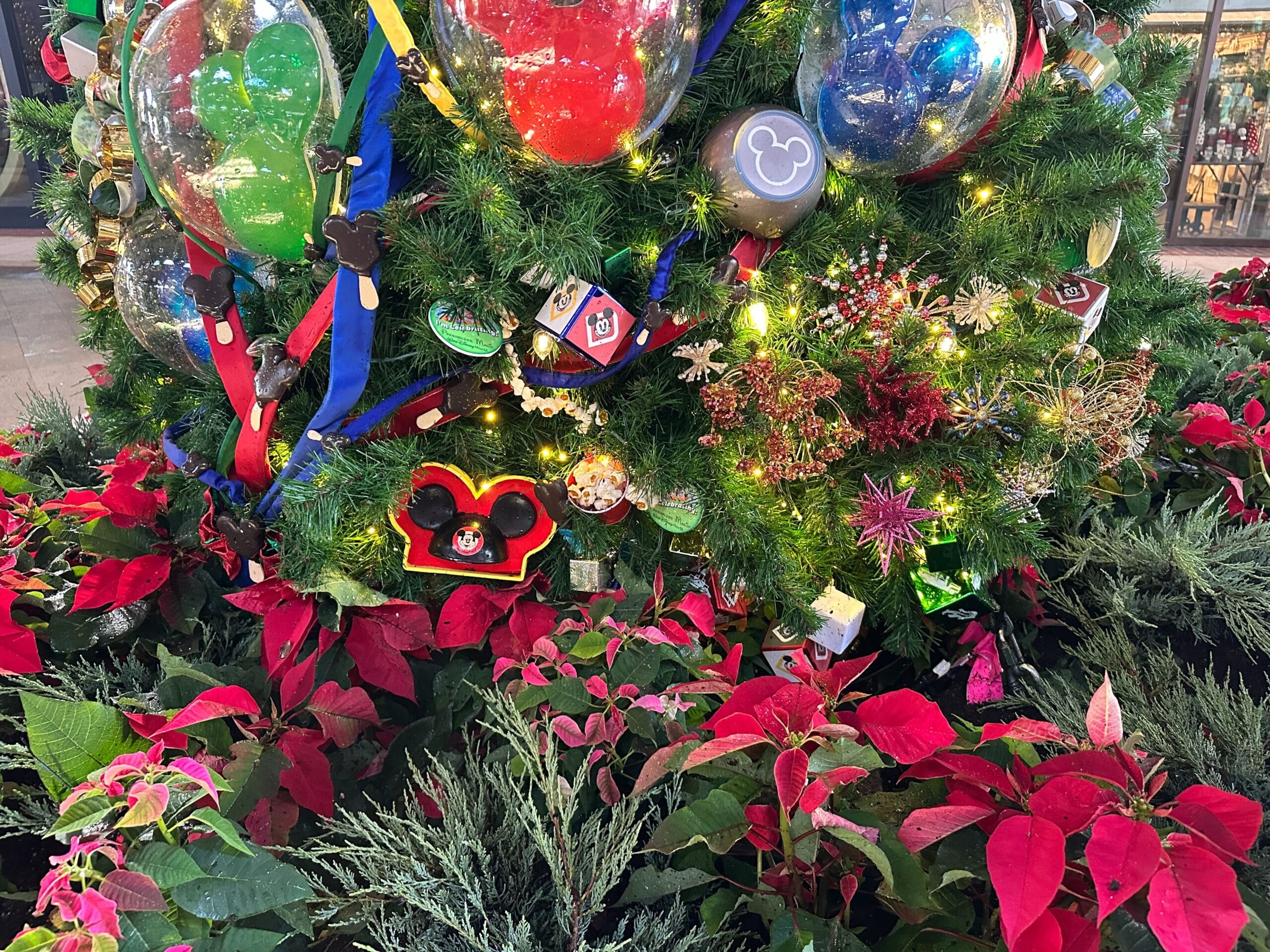 Close-up of a Christmas tree decorated with Disney-themed ornaments and lights, surrounded by red poinsettias and green foliage.