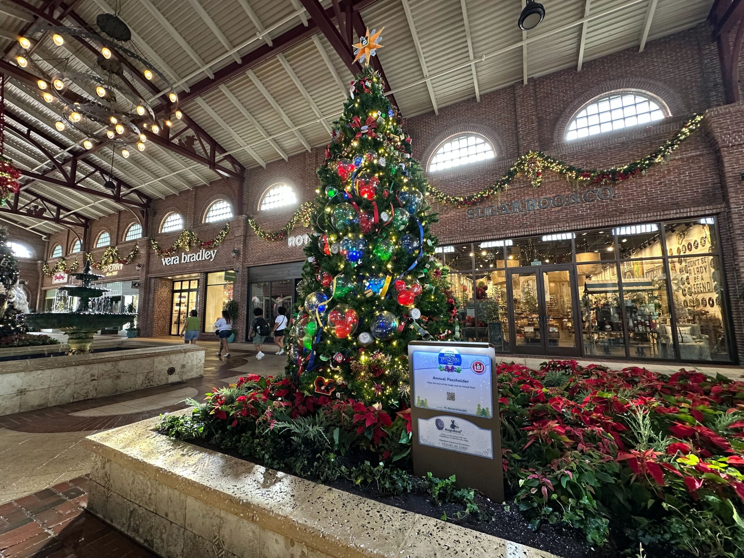 Christmas tree adorned with colorful ornaments stands in a shopping center with brick walls and lighted garlands. Stores in the background include Vera Bradley and Shananigans.
