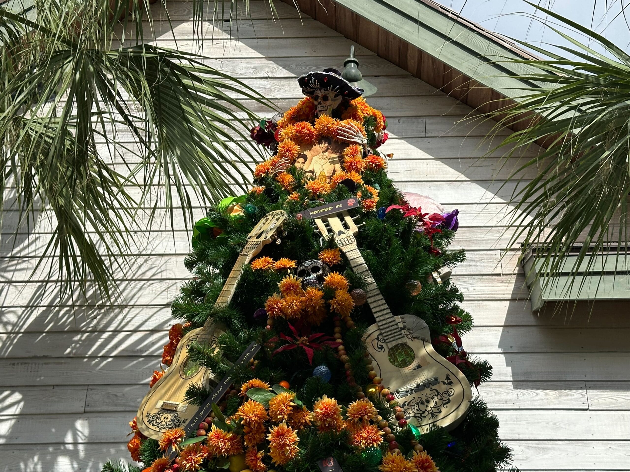 A Christmas tree decorated with guitars, marigold flowers, and a sombrero, set against a wooden wall and surrounded by palm leaves.