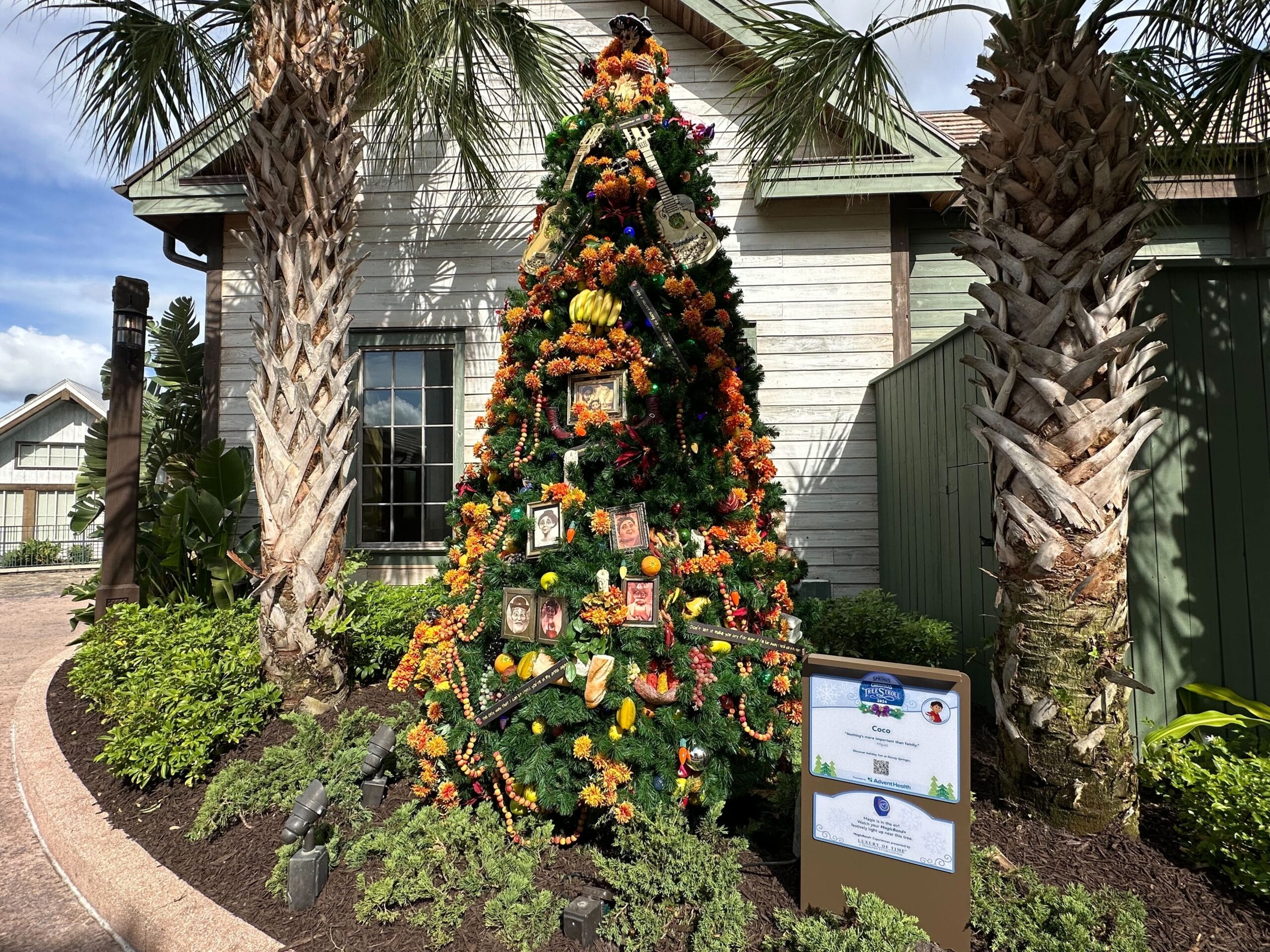 A Christmas tree decorated with animal-themed ornaments stands outside a wooden building, surrounded by palm trees and greenery. A sign is placed in front of the tree.