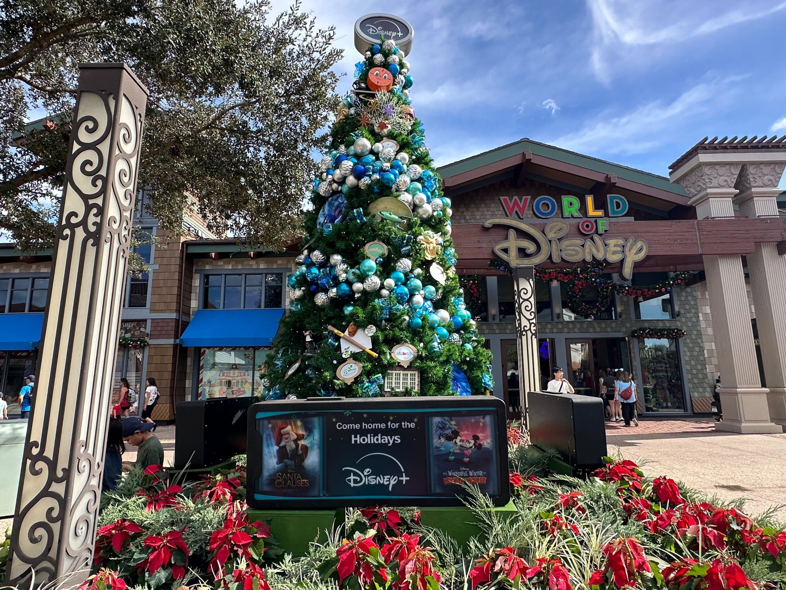 Holiday-themed decorated tree outside the World of Disney store, surrounded by poinsettias and featuring signs for Disney+ shows.