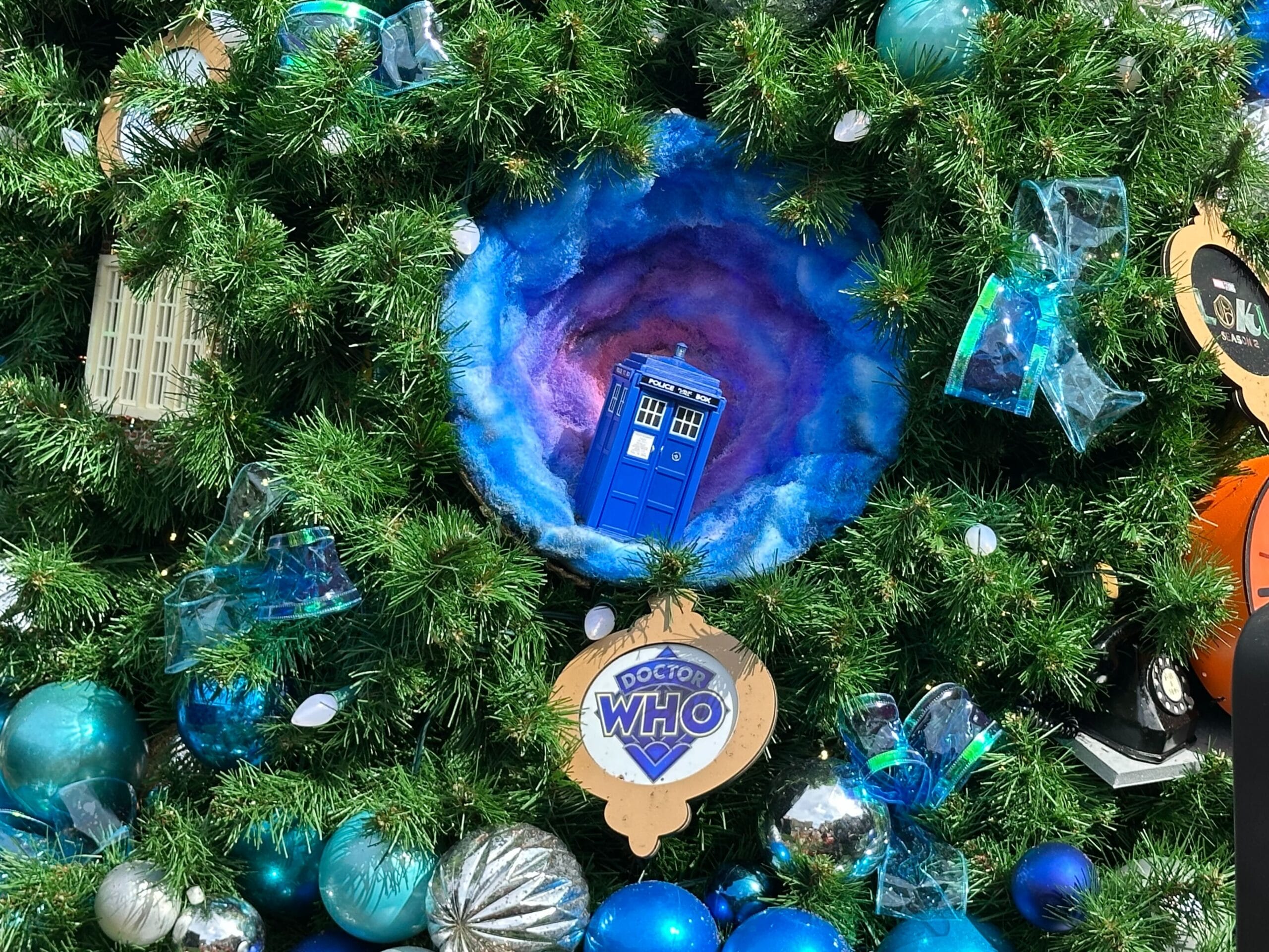A blue TARDIS ornament nestled in green Christmas tree branches, surrounded by blue and silver decorations.
