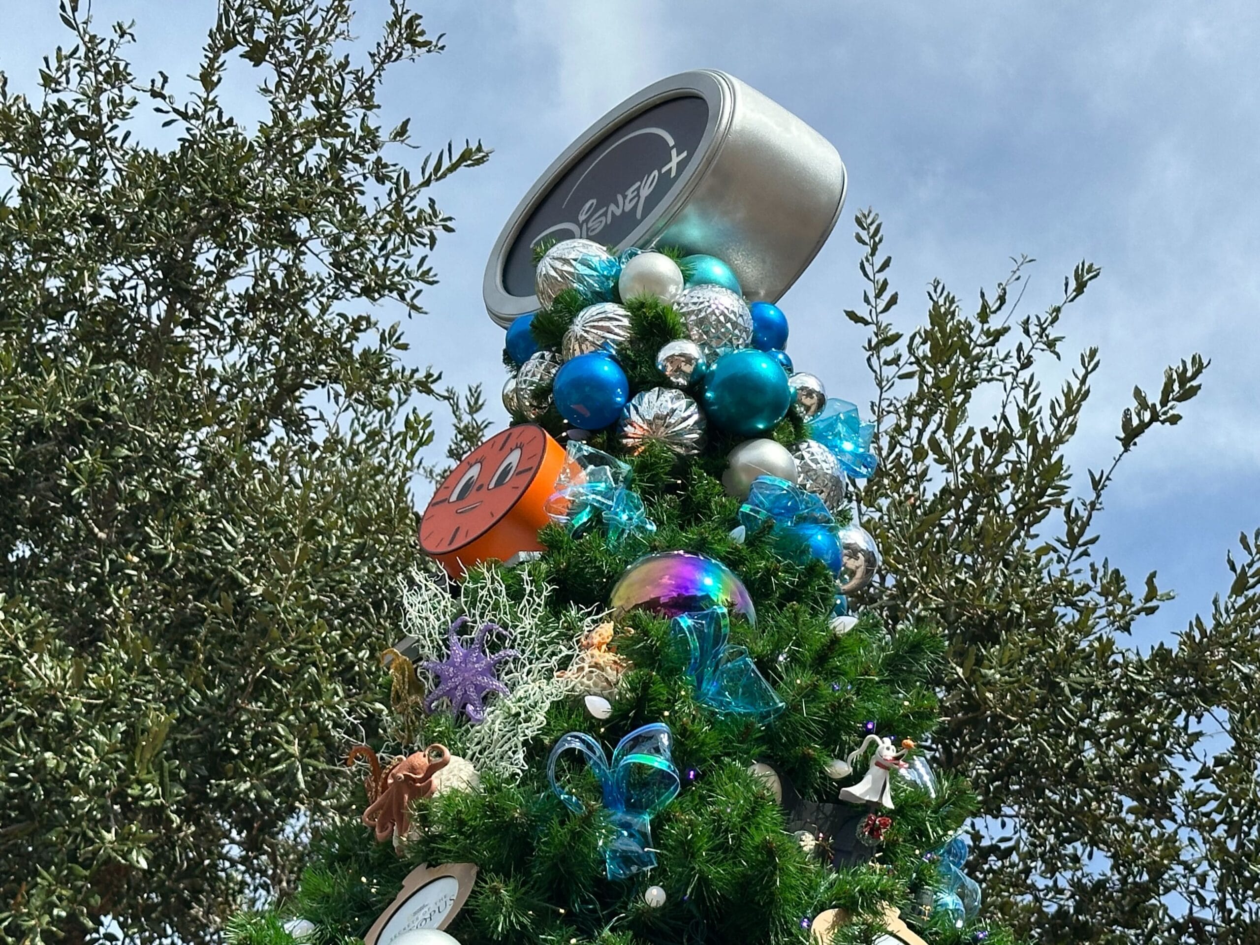 Decorated Christmas tree topped with a cylindrical ornament featuring a logo. Adorned with colorful baubles, bows, and character-themed decorations. Tree is set against a backdrop of trees and sky.