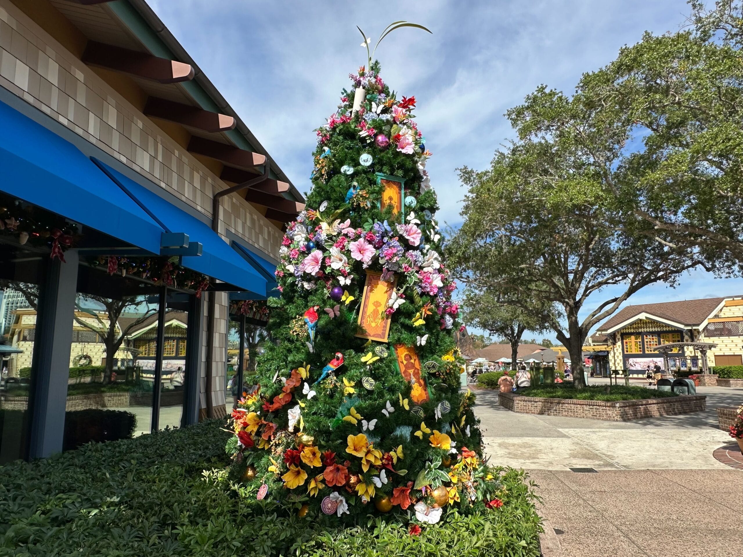 Tall Christmas tree adorned with colorful flowers and portraits, situated outdoors by a blue building under a clear sky.
