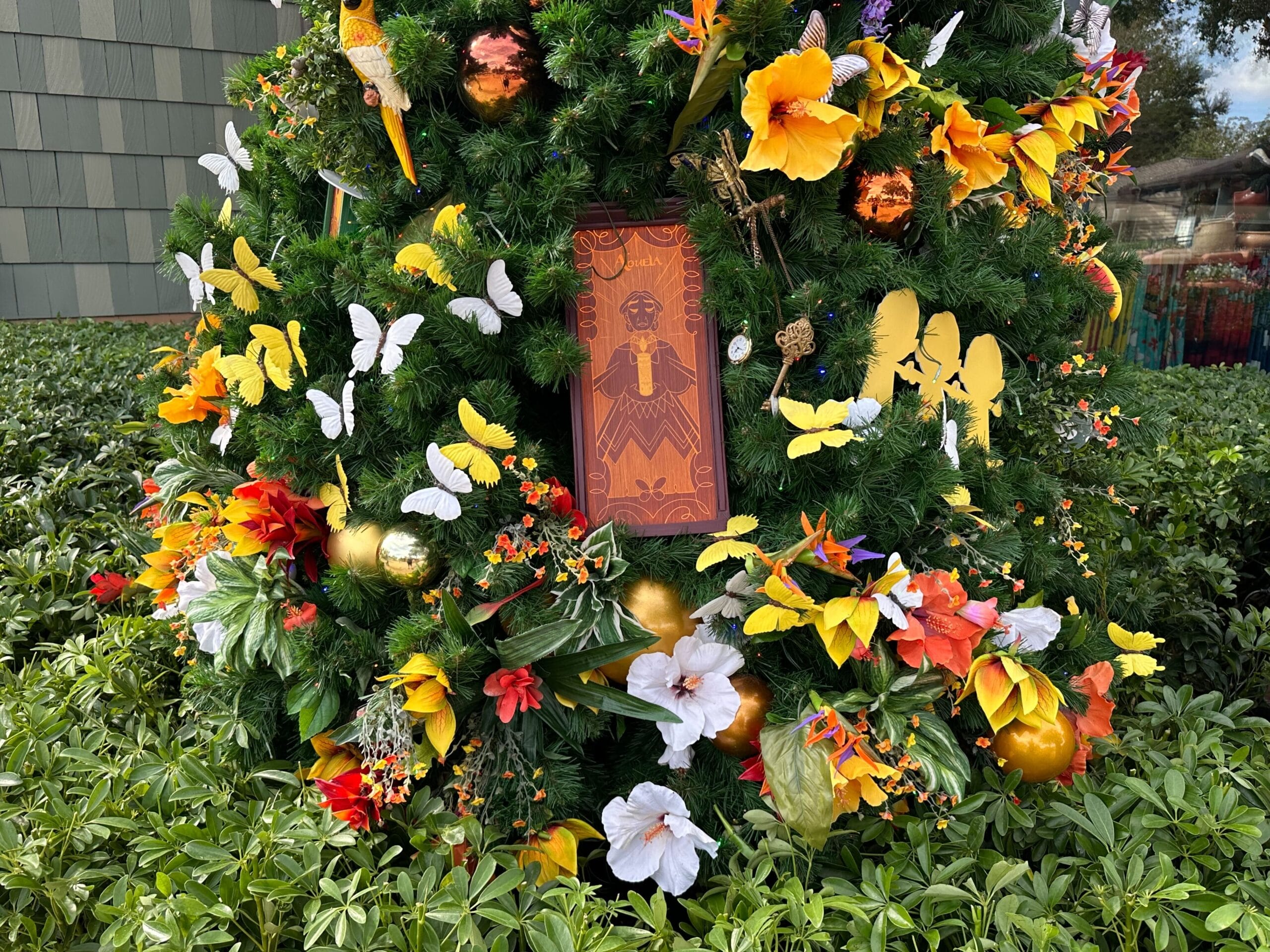 A decorative wreath adorned with colorful flowers, butterflies, and gold ornaments, featuring a central rectangular artwork. The wreath is set amidst green foliage.