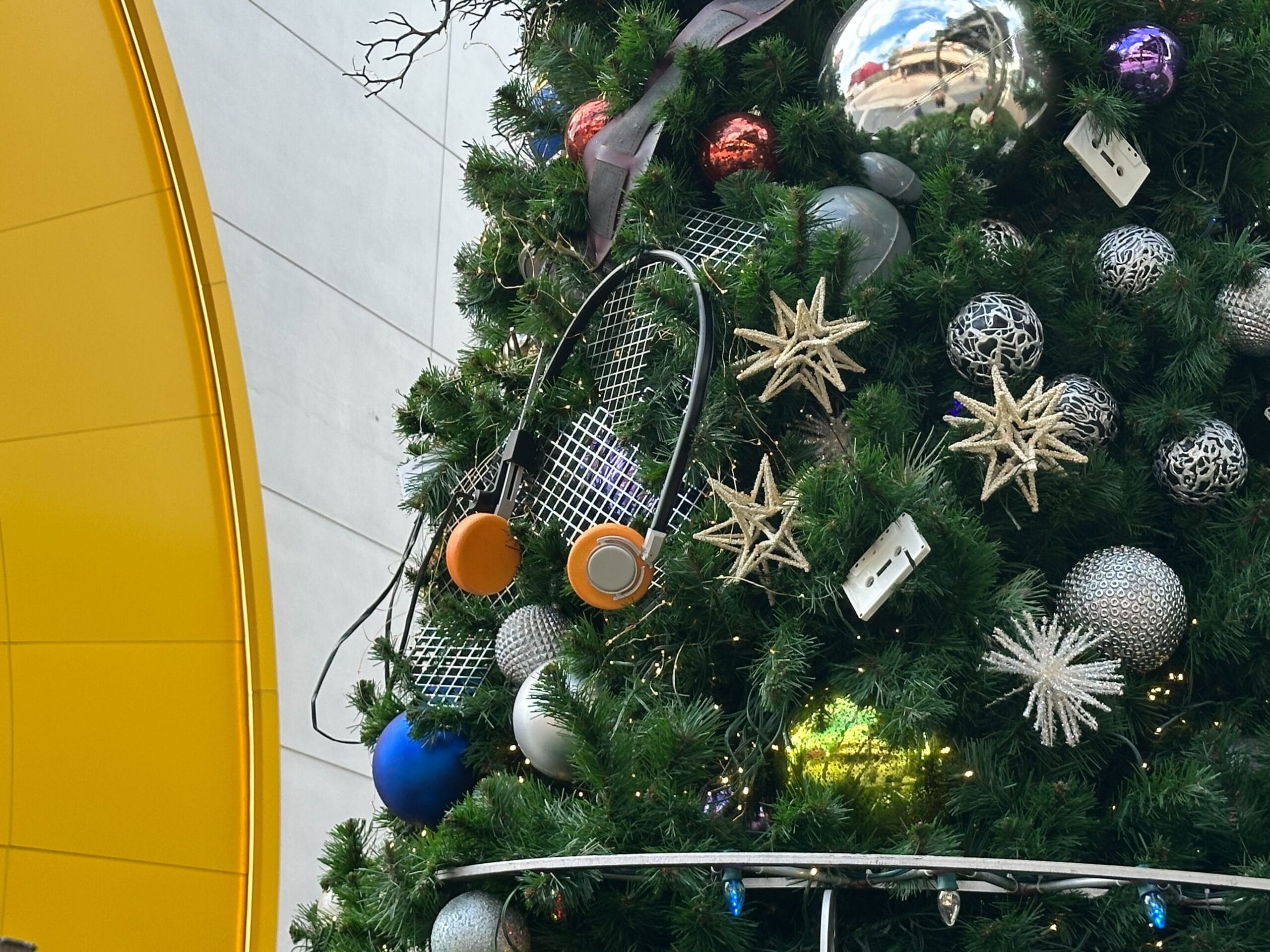 Close-up of a decorated Christmas tree with shiny ornaments, star-shaped decorations, reflective spheres, and a yellow wall in the background.