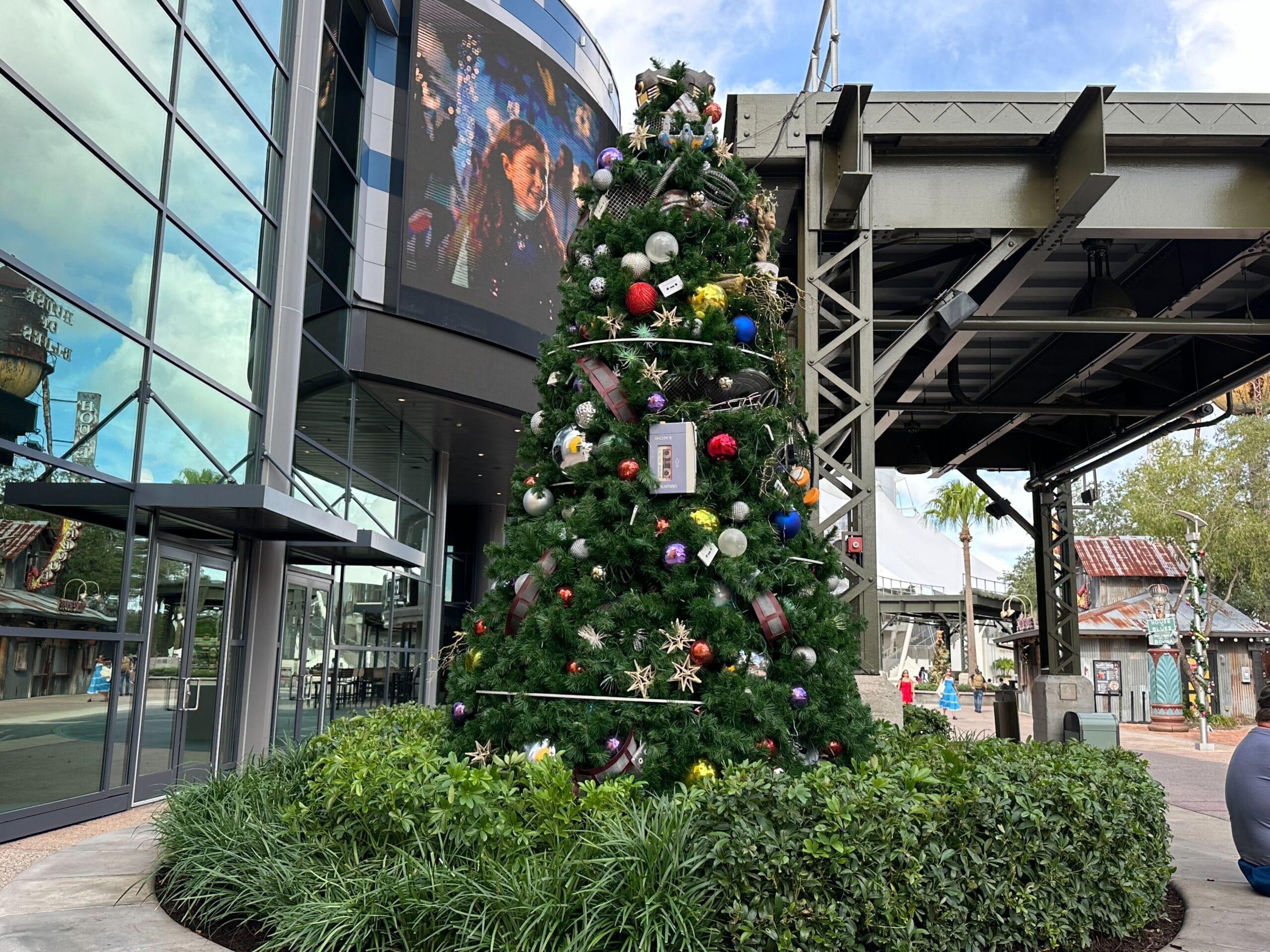 A decorated Christmas tree stands outside a modern building under a clear sky, surrounded by greenery and metal structures.