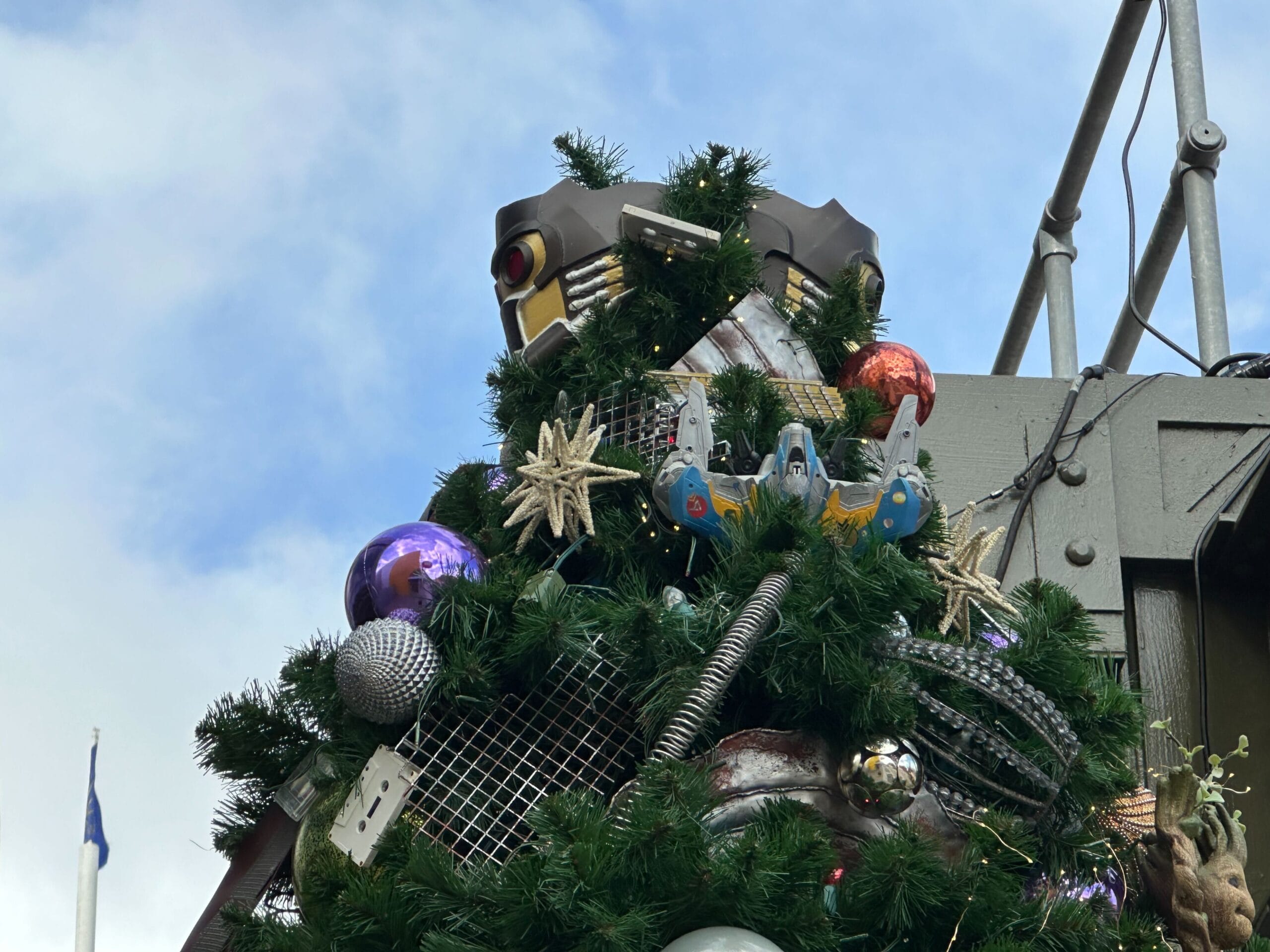 Festive tree decorated with colorful ornaments, a toy spaceship, and a large robot head on top against a blue sky backdrop.
