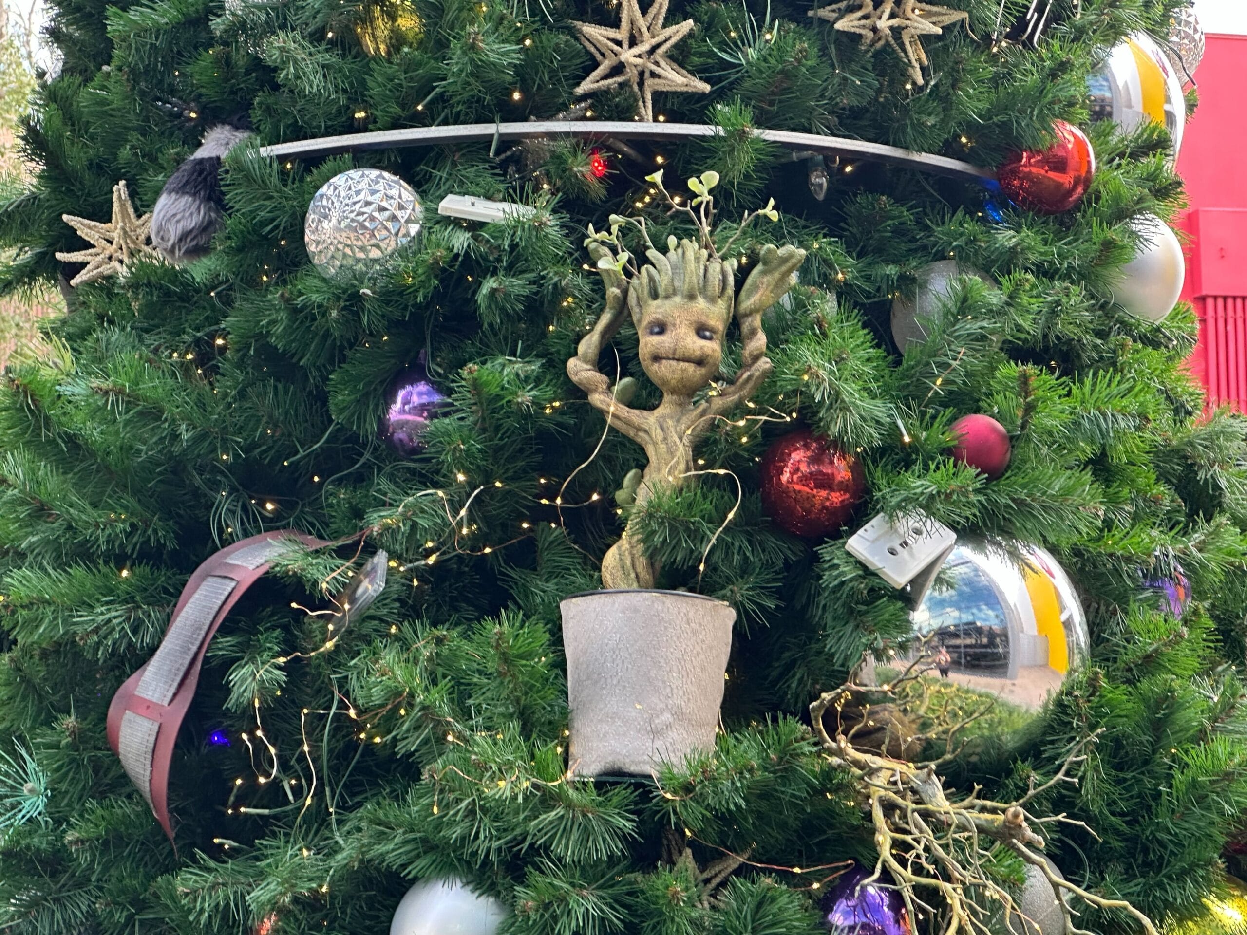 A Christmas tree decorated with red, silver, and gold ornaments features a potted Groot figure surrounded by lights.