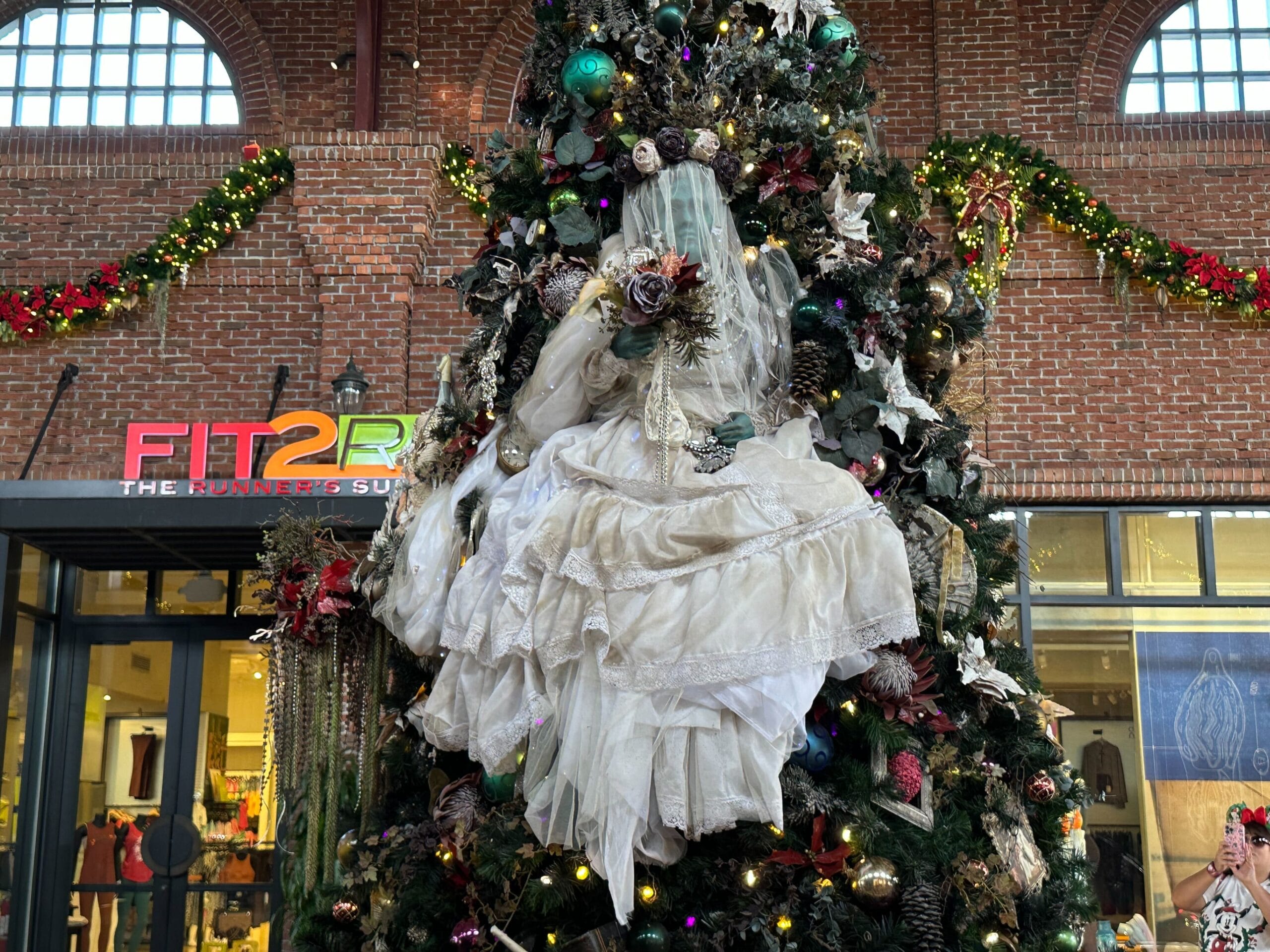 A mannequin dressed as a ghostly bride sits in a large, decorated Christmas tree inside a building, with a "FIT2RUN" store in the background.