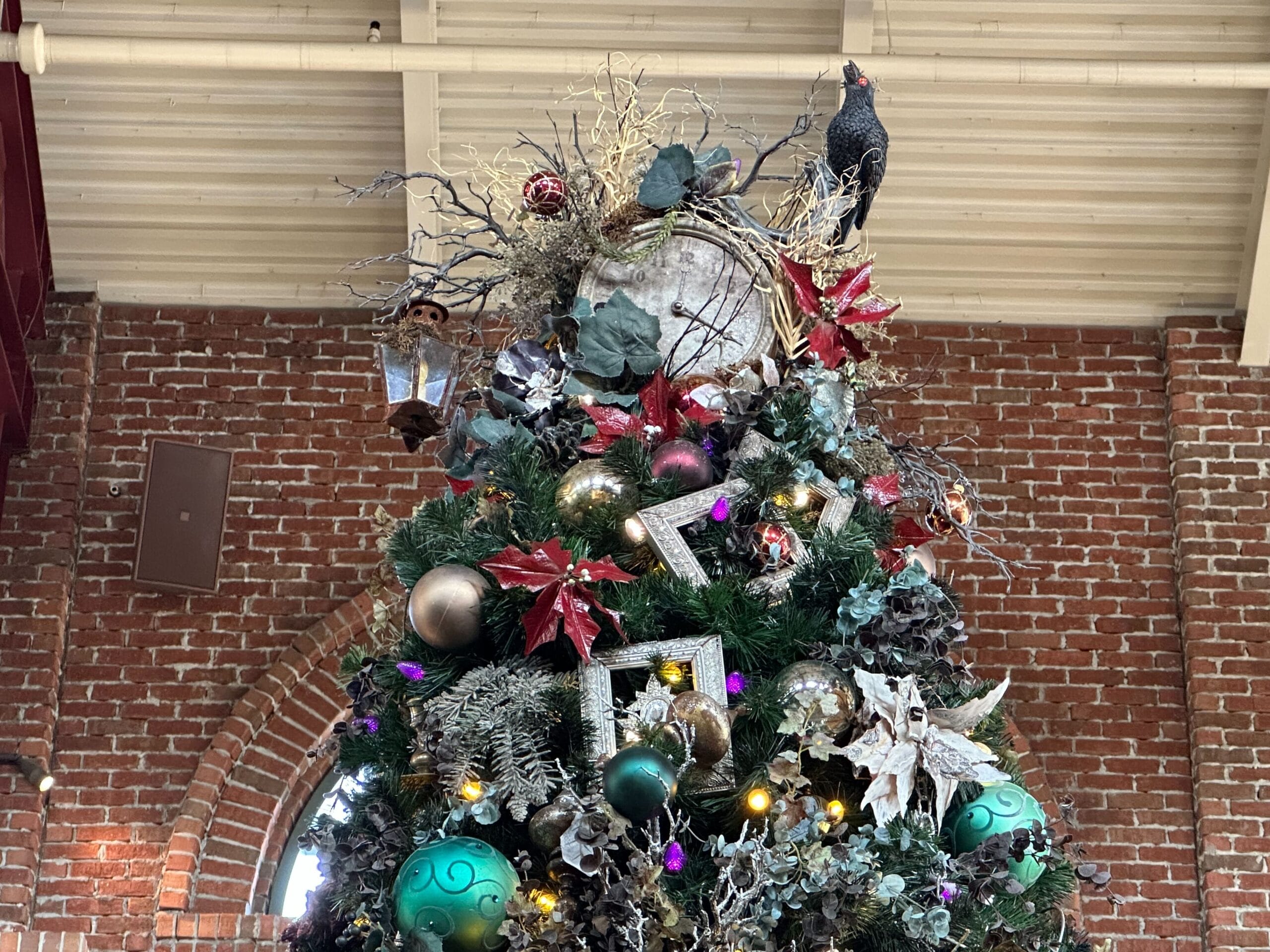 Ornately decorated Christmas tree with red, green, and gold ornaments, greenery, baubles, and a bird figure on top, set against a brick wall background.