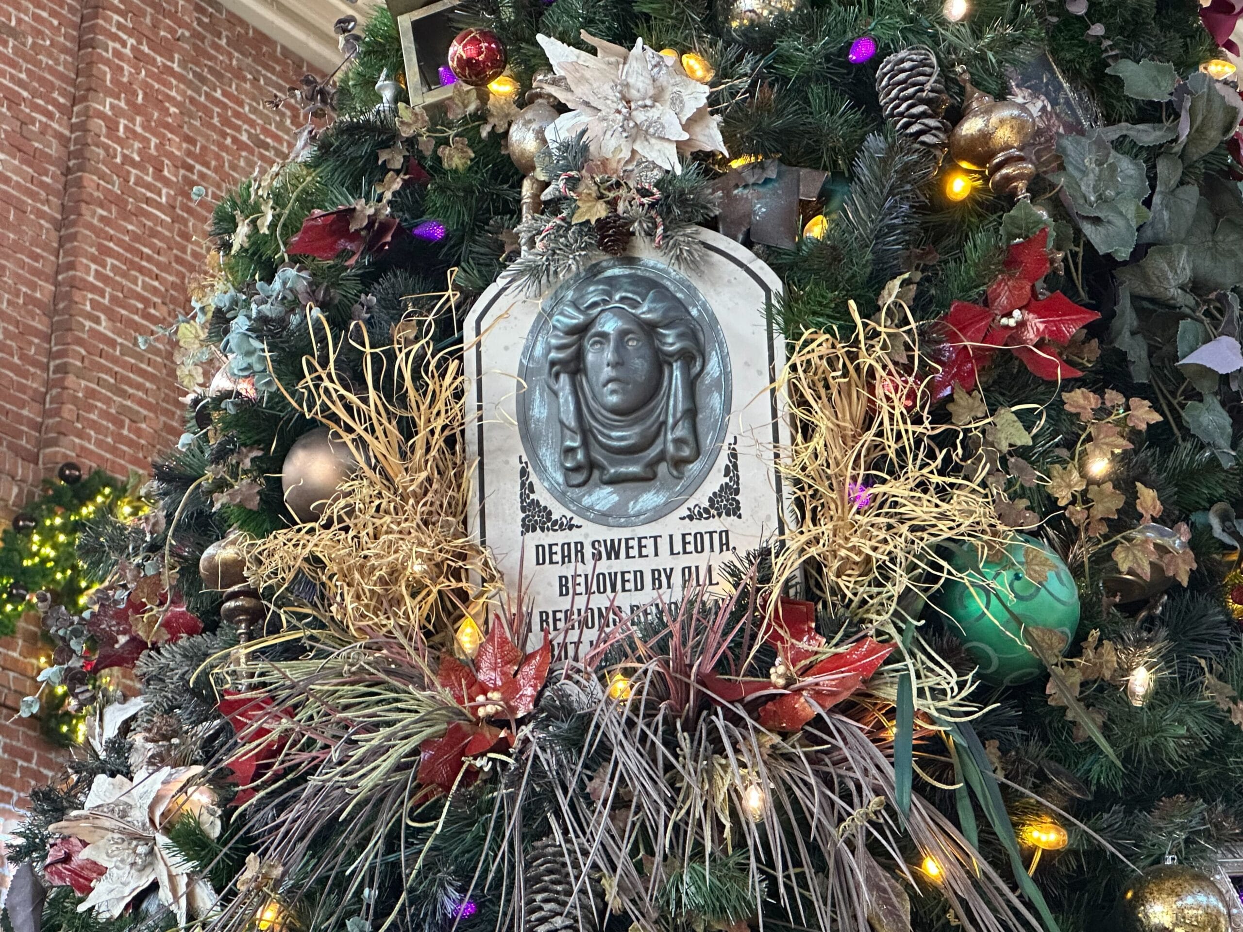 Decorated Christmas tree with a plaque featuring a face and the words "Dear Sweet Leota, Beloved by all, in regions beyond now, but having a ball." surrounded by ornaments and leaves.