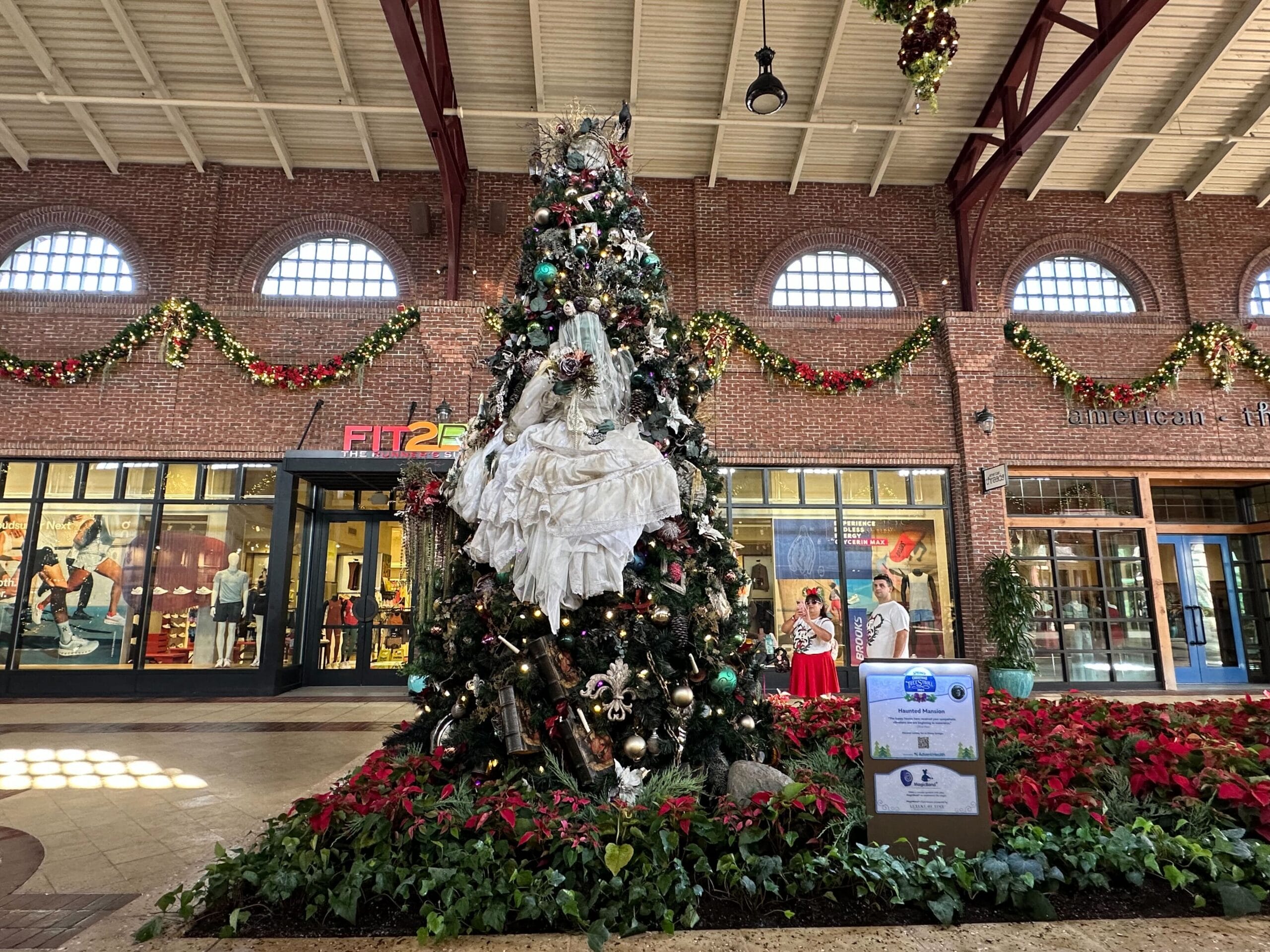 A decorated Christmas tree stands in a mall, surrounded by poinsettias. Holiday garlands are hung on the walls.