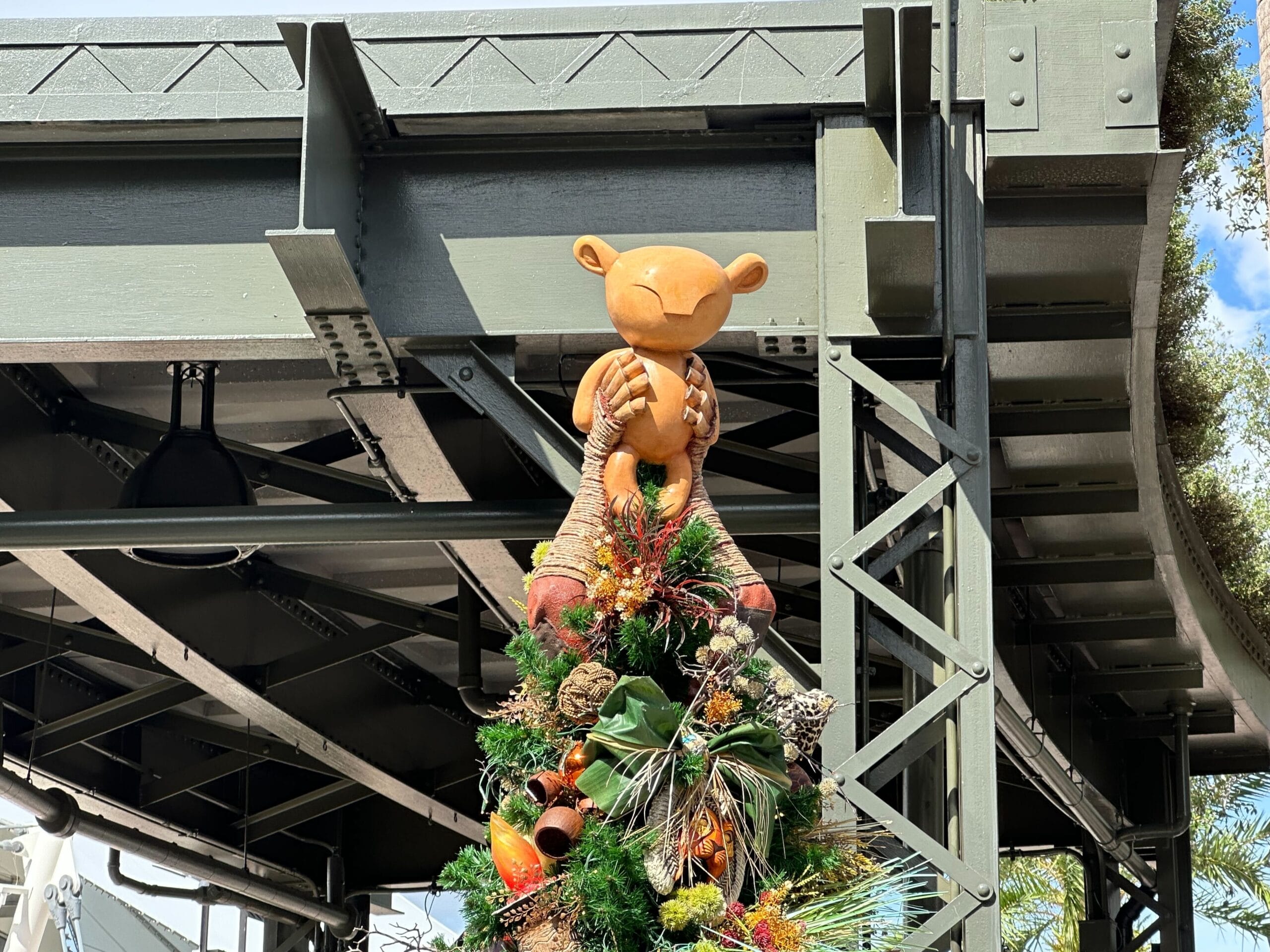 A decorative figure sits atop a Christmas tree under a metal bridge, surrounded by greenery and holiday ornaments.