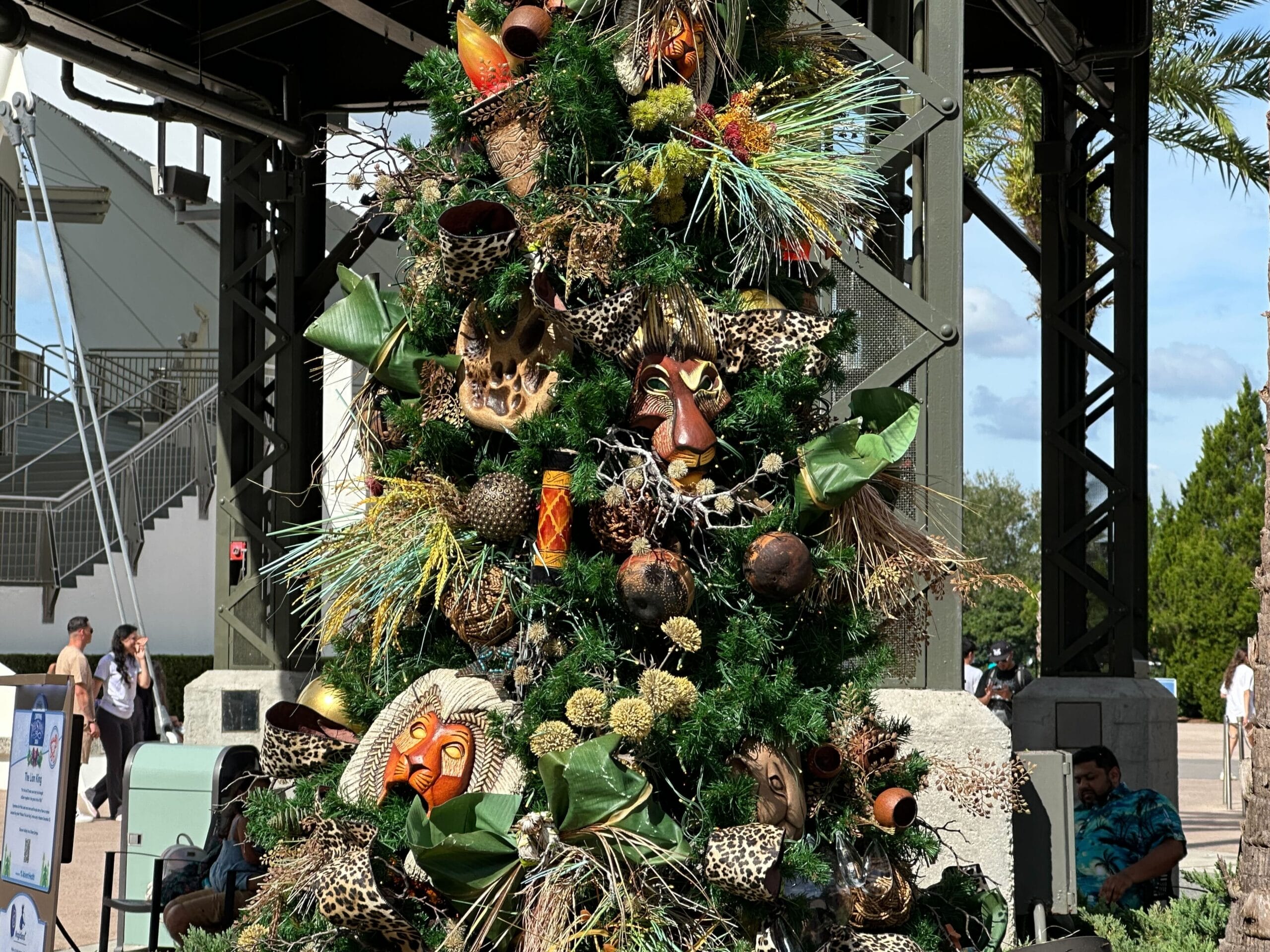 A Christmas tree decorated with African-themed masks, animal print fabrics, and tropical foliage stands outdoors under a metal structure.
