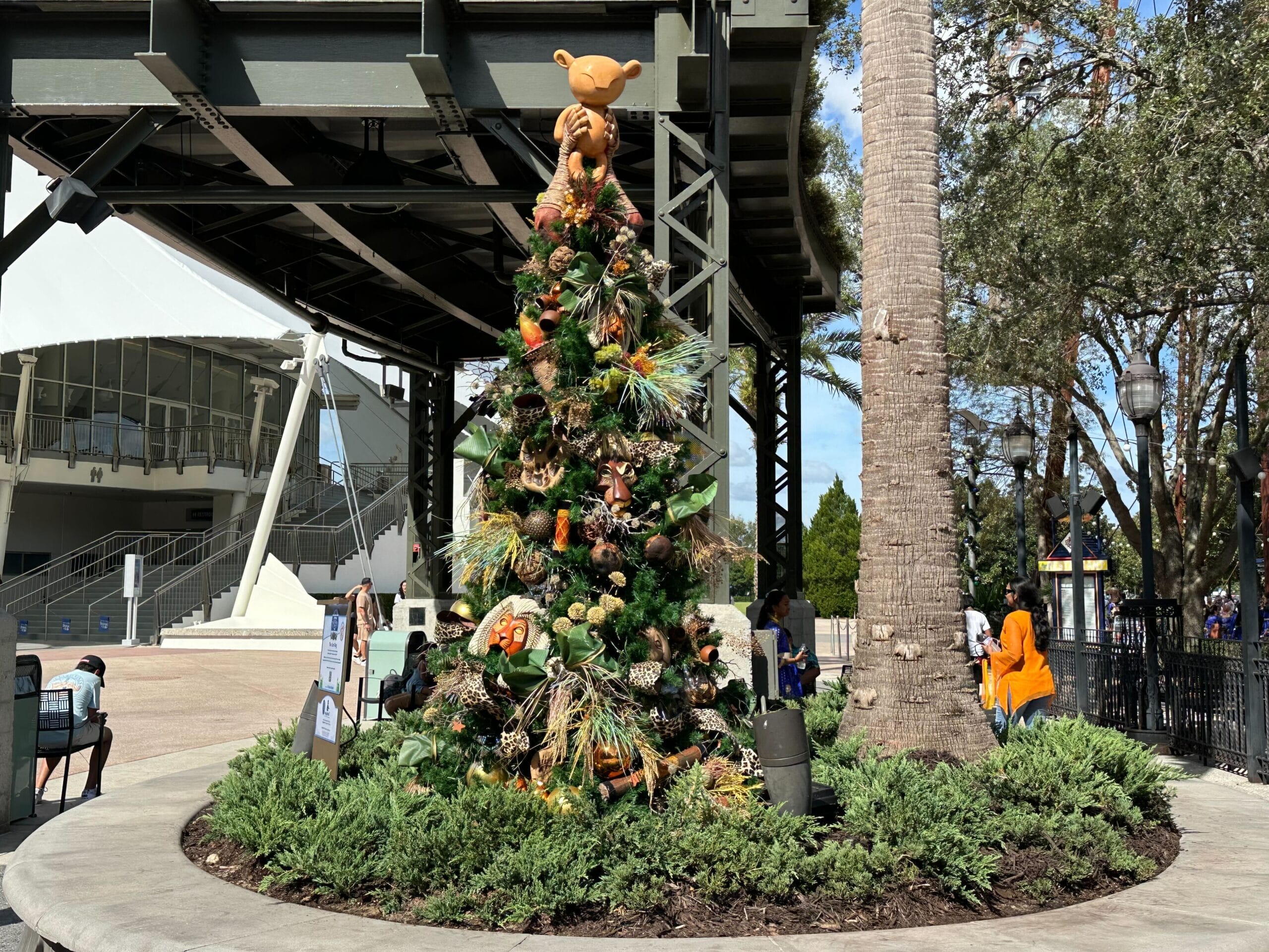Christmas tree adorned with colorful decorations, under an outdoor structure, with people sitting nearby and trees in the background.
