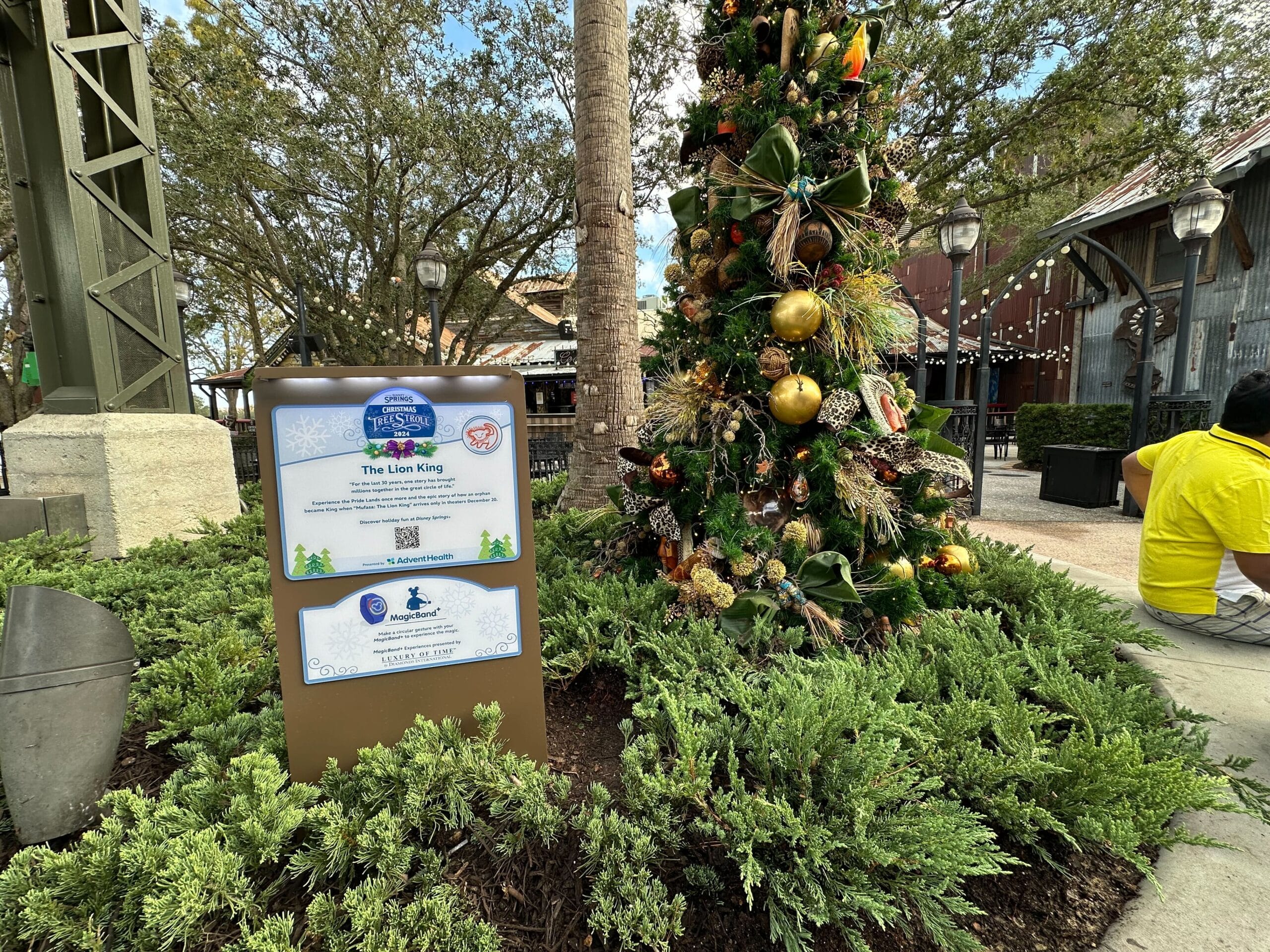 Christmas tree decorated with themed ornaments near a sign about "The Lion King," surrounded by greenery and outdoor lamps in a park setting.