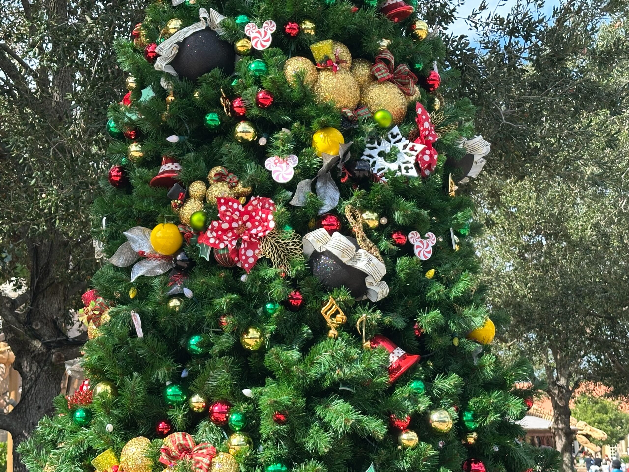 A decorated Christmas tree with green, red, and gold ornaments, large bows, and various festive decorations, set outdoors with trees in the background.