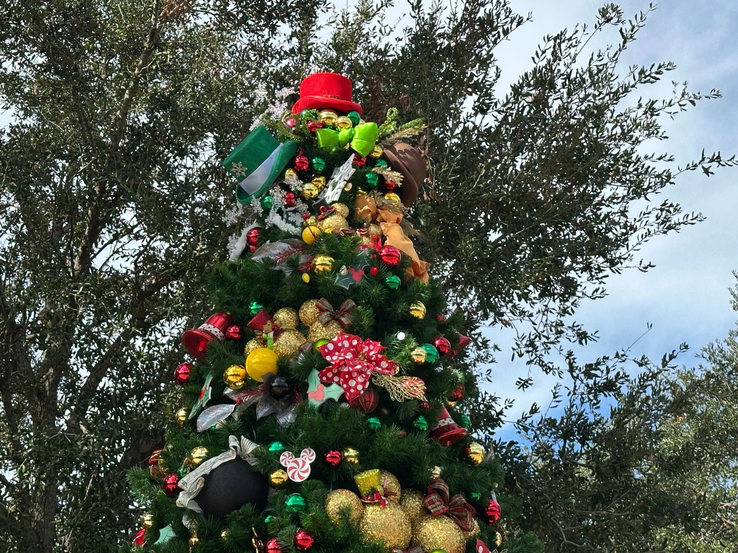 Festive Christmas tree adorned with various decorations, including a red hat, bows, gifts, and shiny ornaments, set against a backdrop of green trees and a blue sky.
