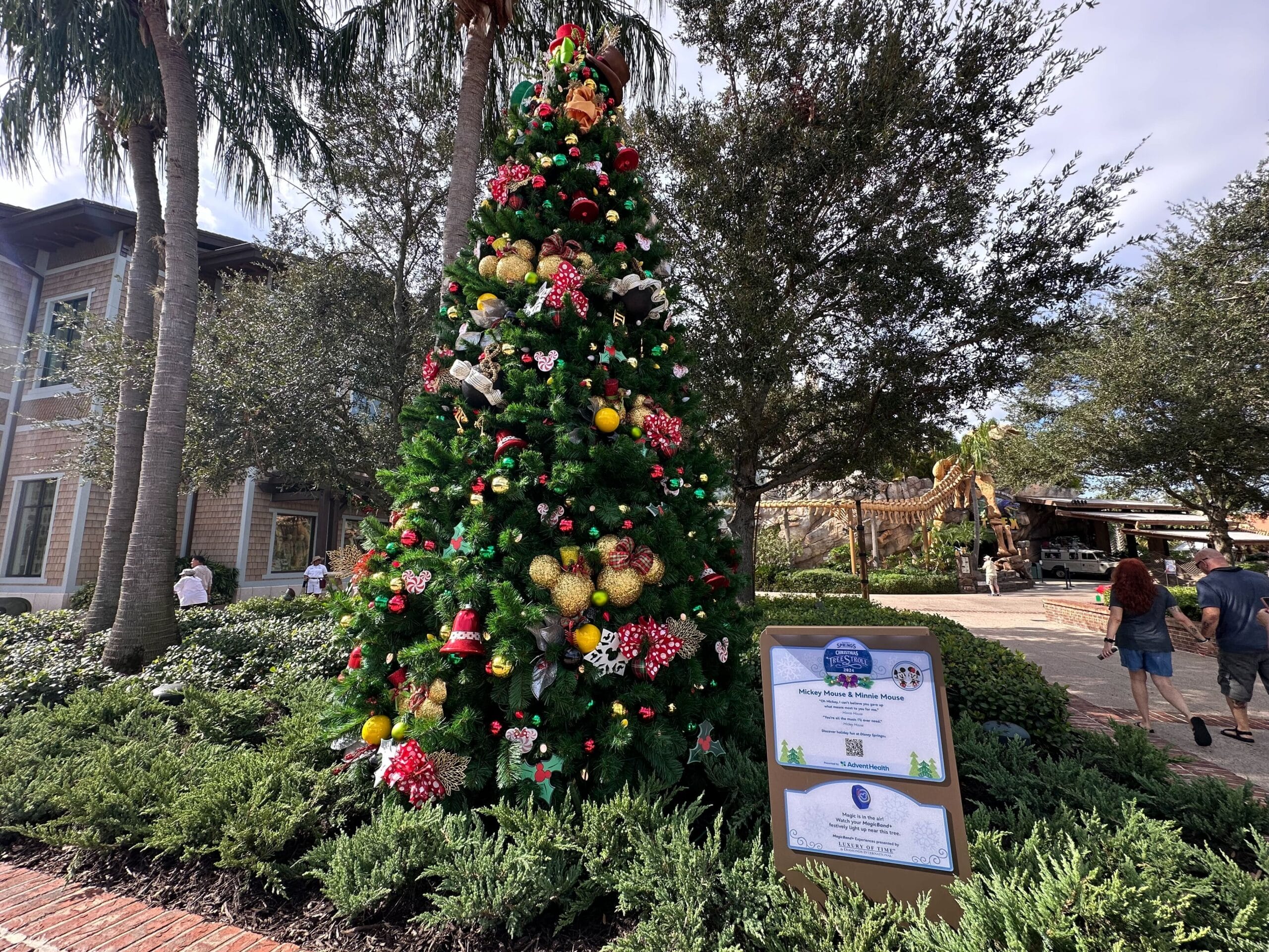 A large decorated Christmas tree stands outdoors surrounded by greenery and trees. A sign with event information is placed in front of the tree. People walk nearby.