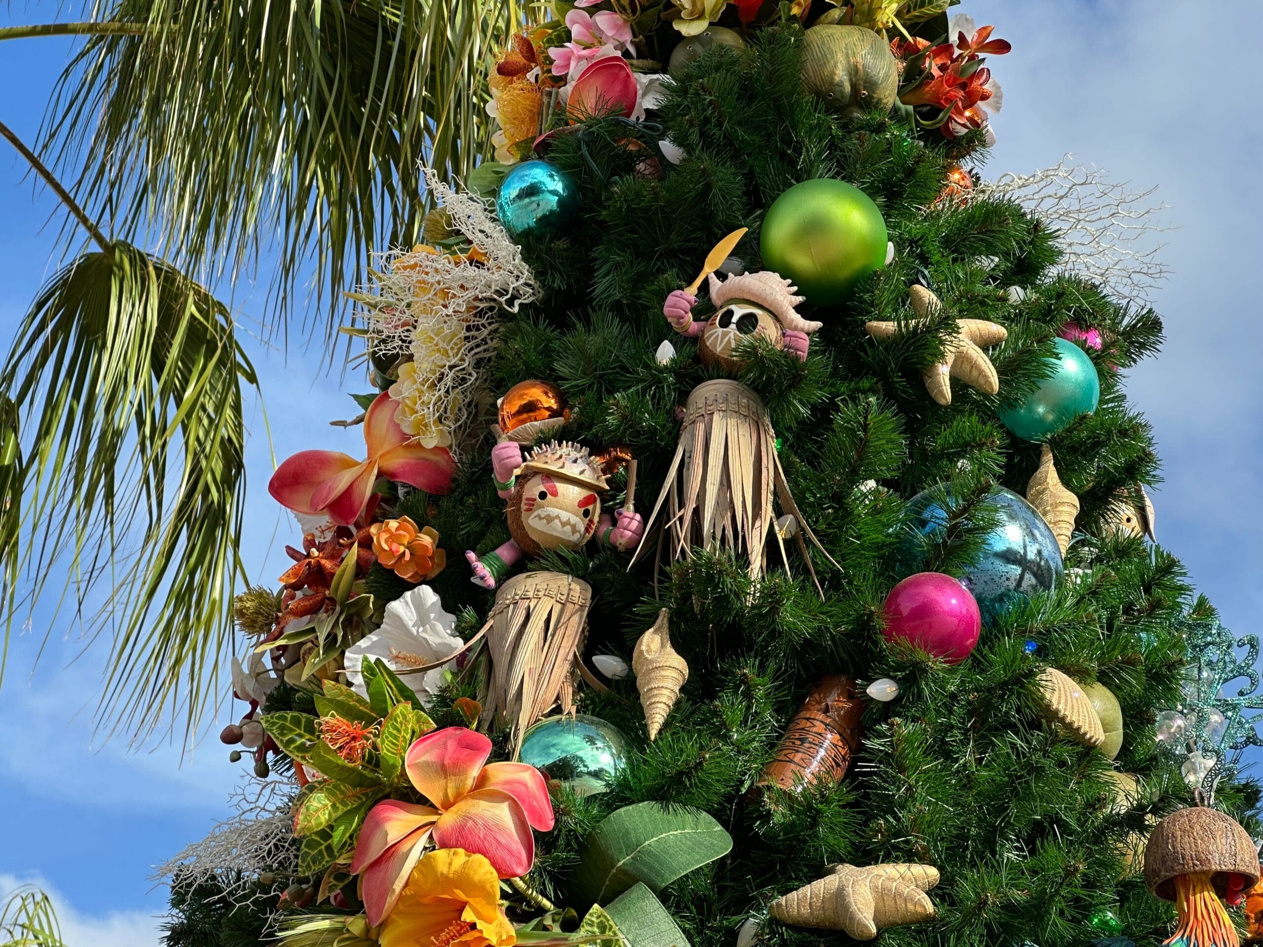 Decorated Christmas tree with tropical ornaments, including tiki figures, colorful baubles, flowers, and seashells, under palm branches against a blue sky.
