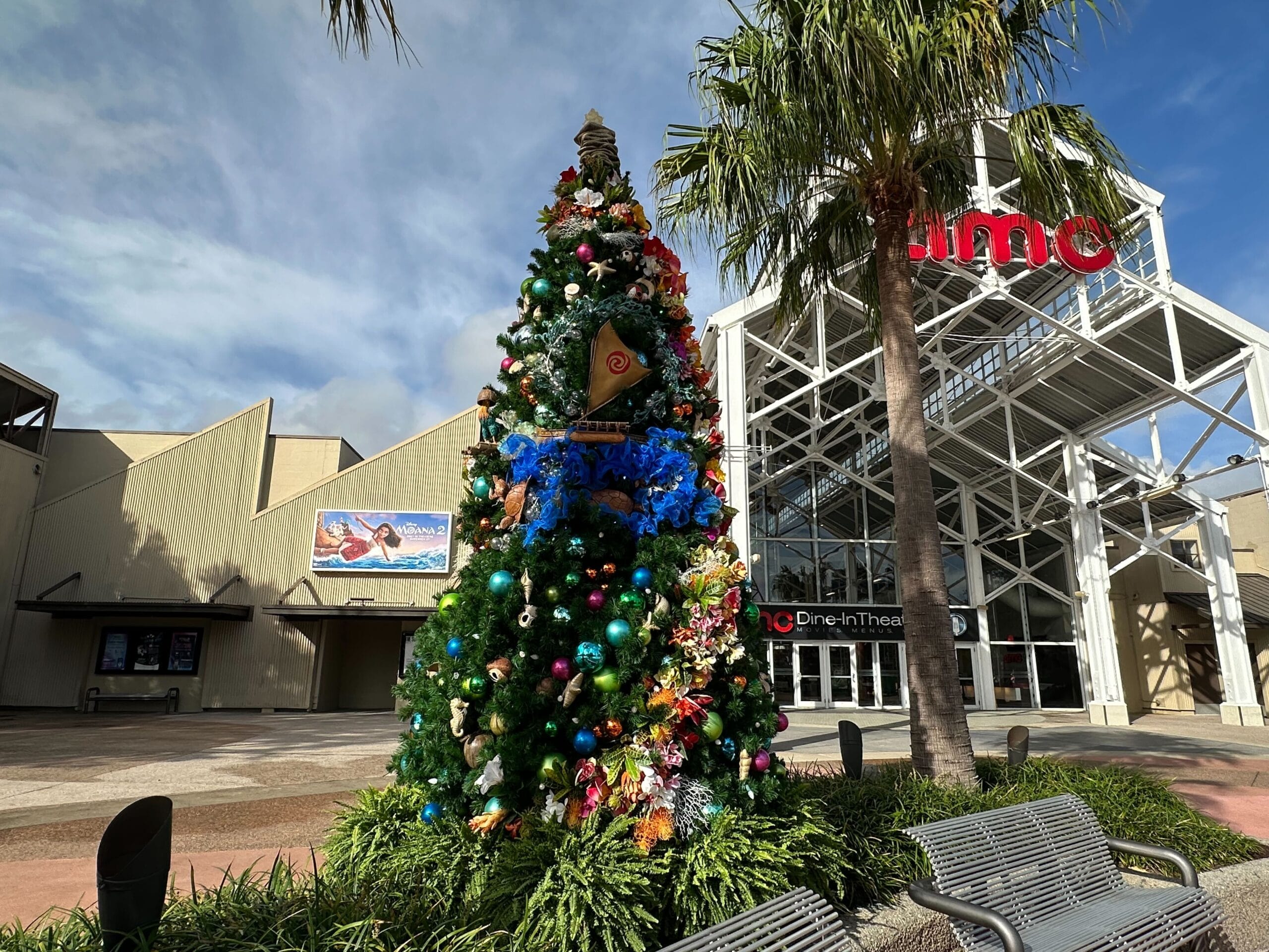 A decorated Christmas tree with colorful ornaments stands outside a cinema building under a clear sky.