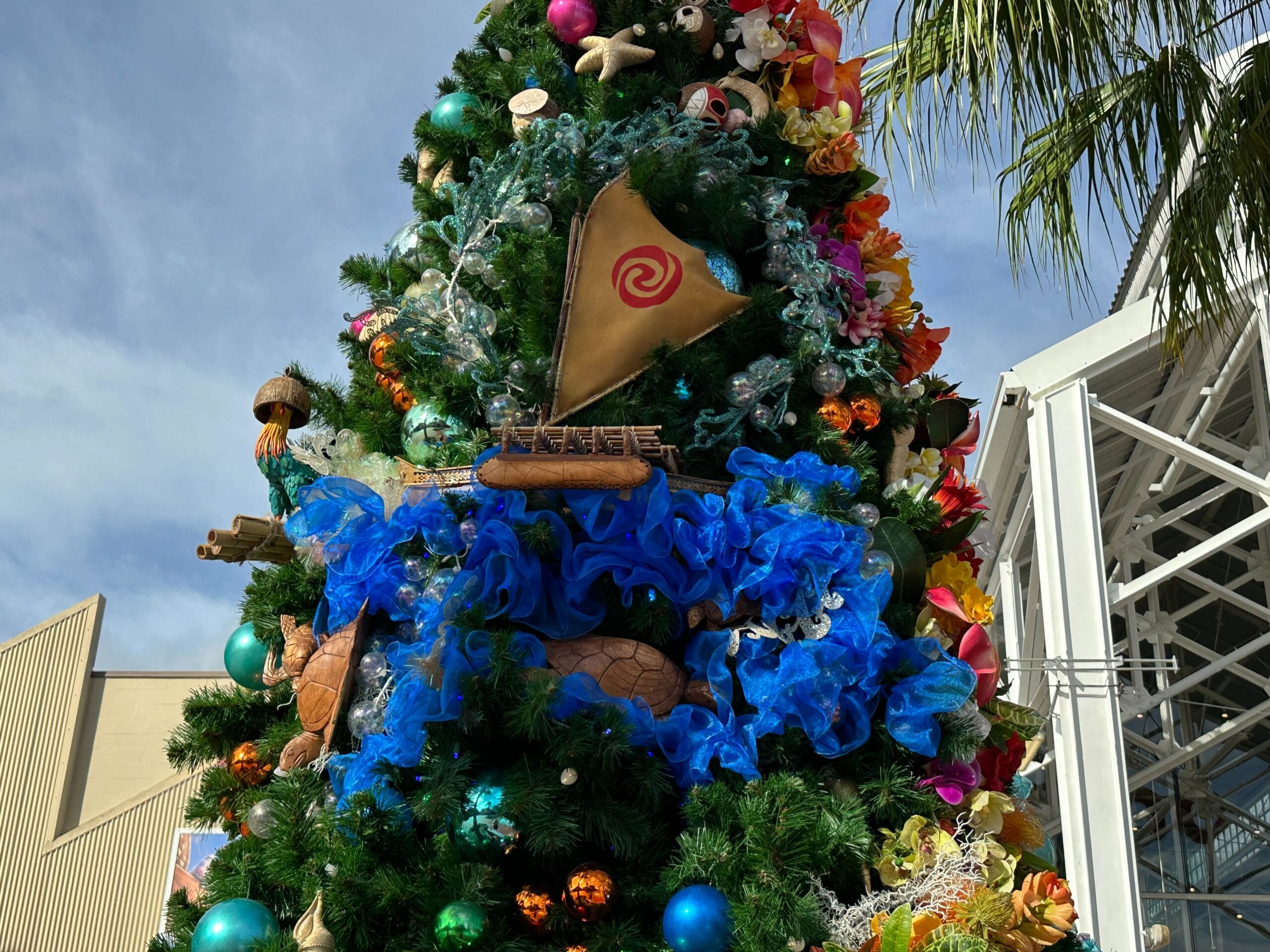 A decorated Christmas tree with a tropical theme, featuring a small sailboat ornament and various ocean-themed decorations, set against a blue sky and partial building structure.
