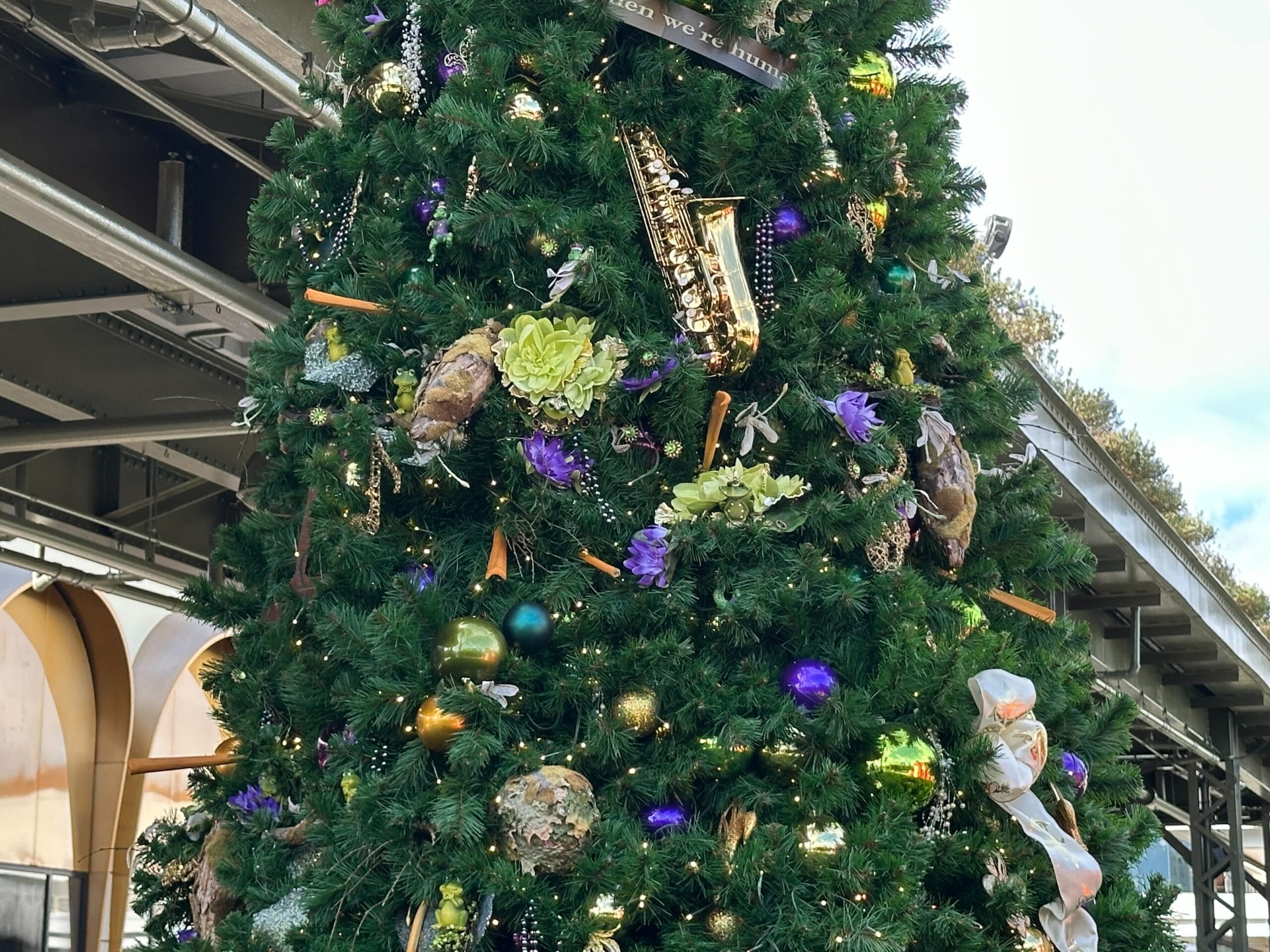 A decorated Christmas tree with ornaments, including a saxophone, flowers, and colorful baubles, stands under a metal structure.