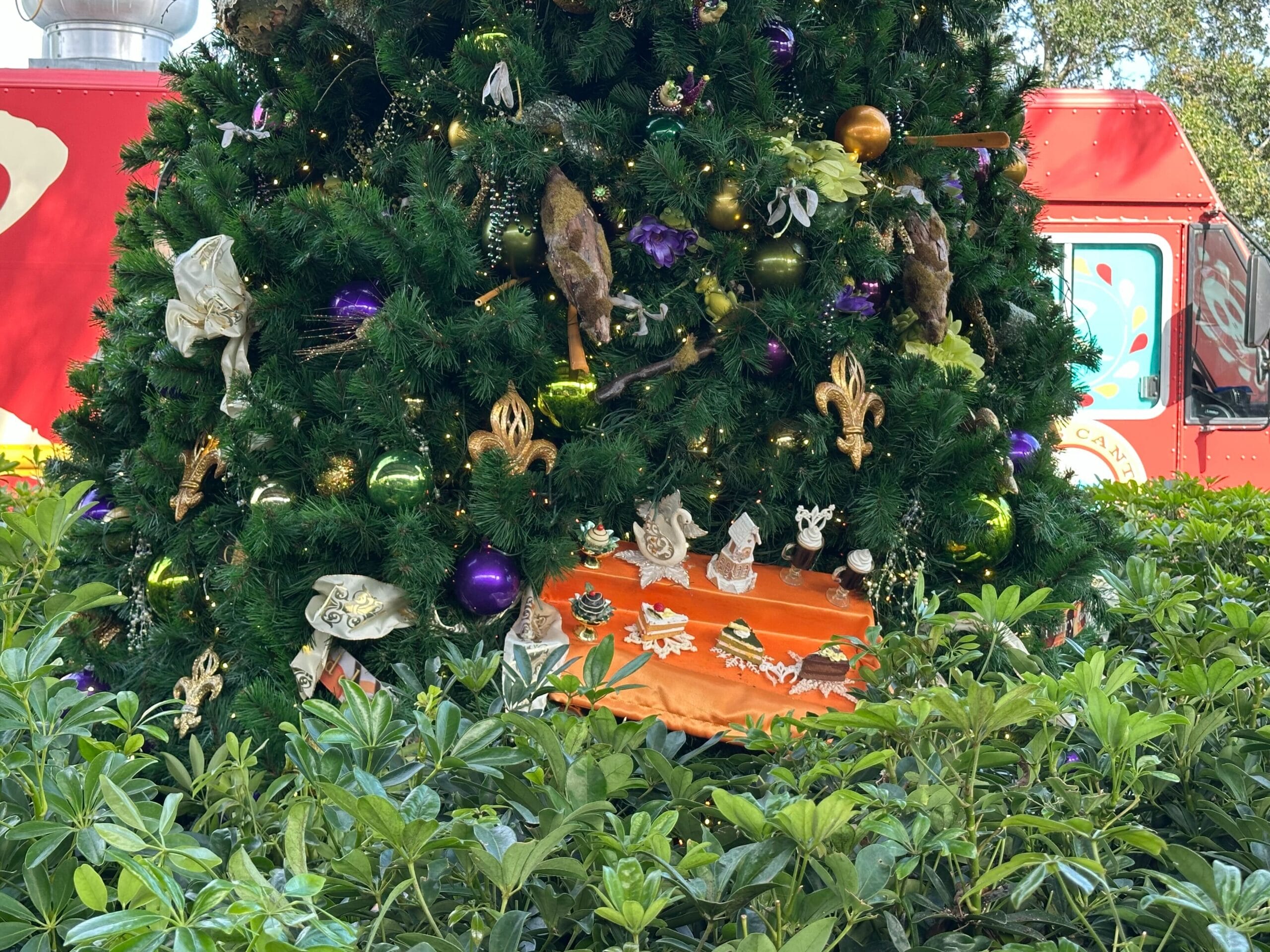Decorated Christmas tree with purple and gold ornaments, surrounded by green shrubs. A red vehicle is partially visible in the background.