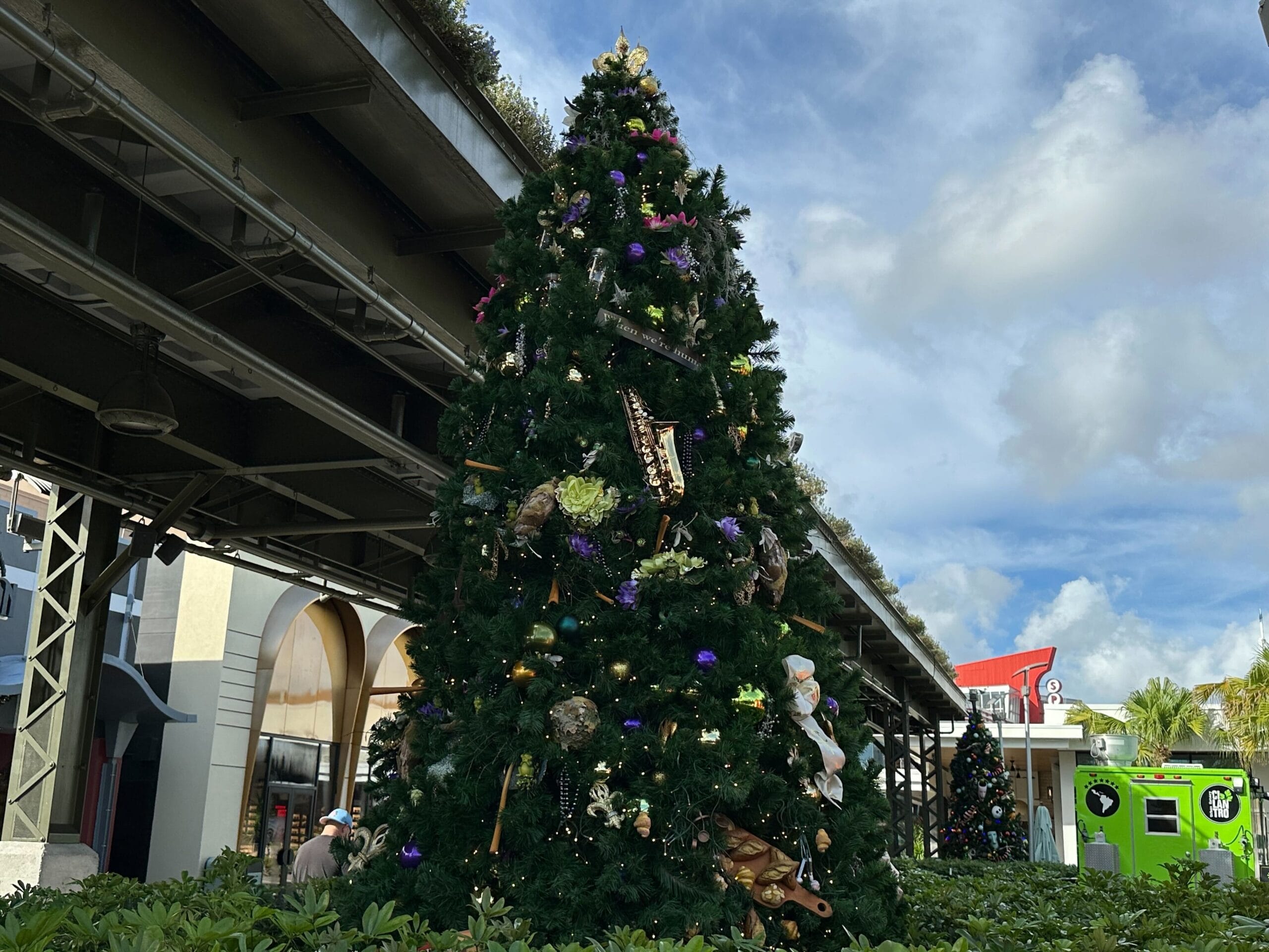 Tall Christmas tree adorned with various decorations, set outdoors under a partly cloudy sky, with nearby buildings and greenery.