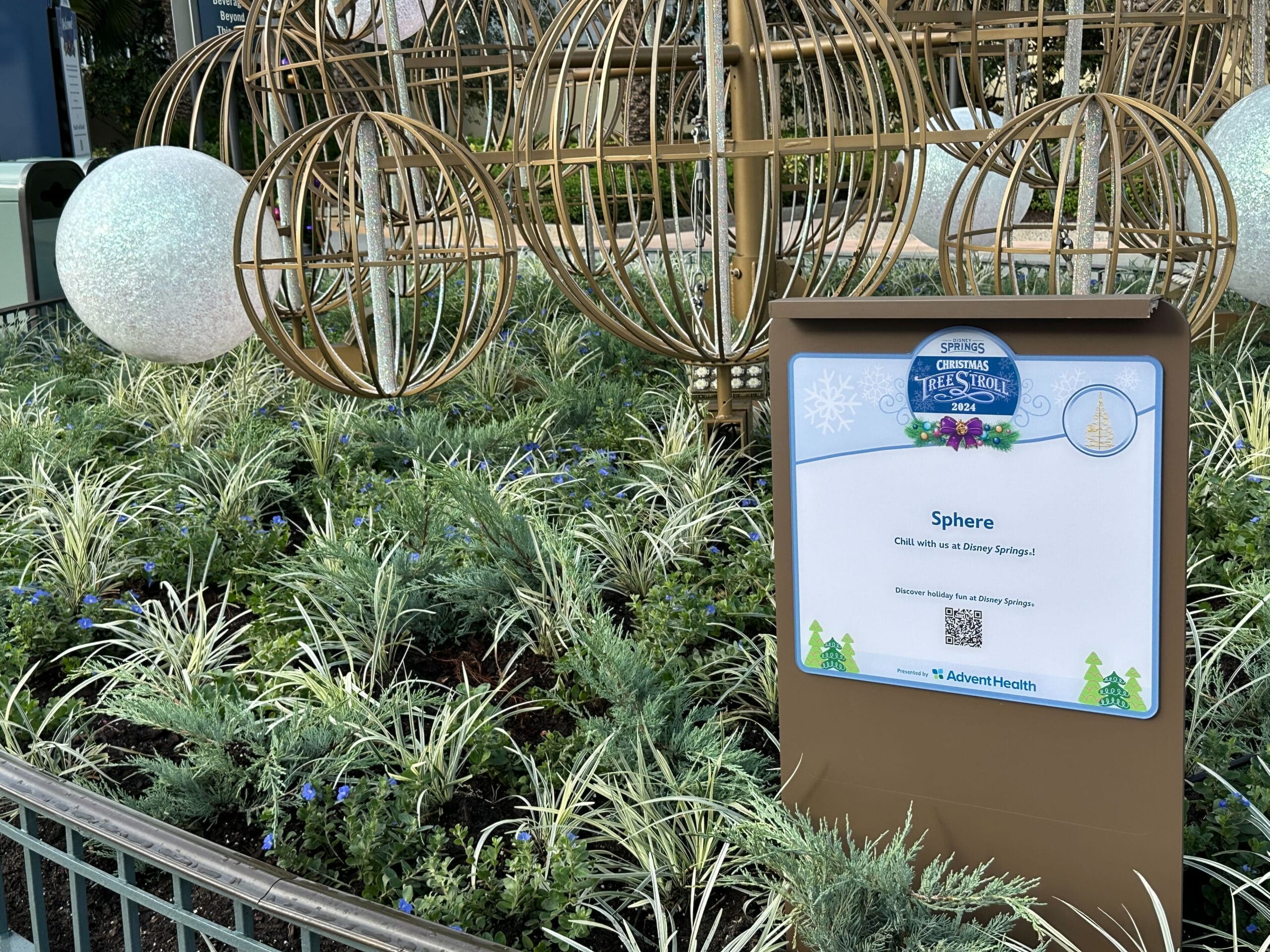 A garden display with decorative metal spheres and plants. A sign labeled "Sphere" describes the installation at Disney Springs and credits AdventHealth.